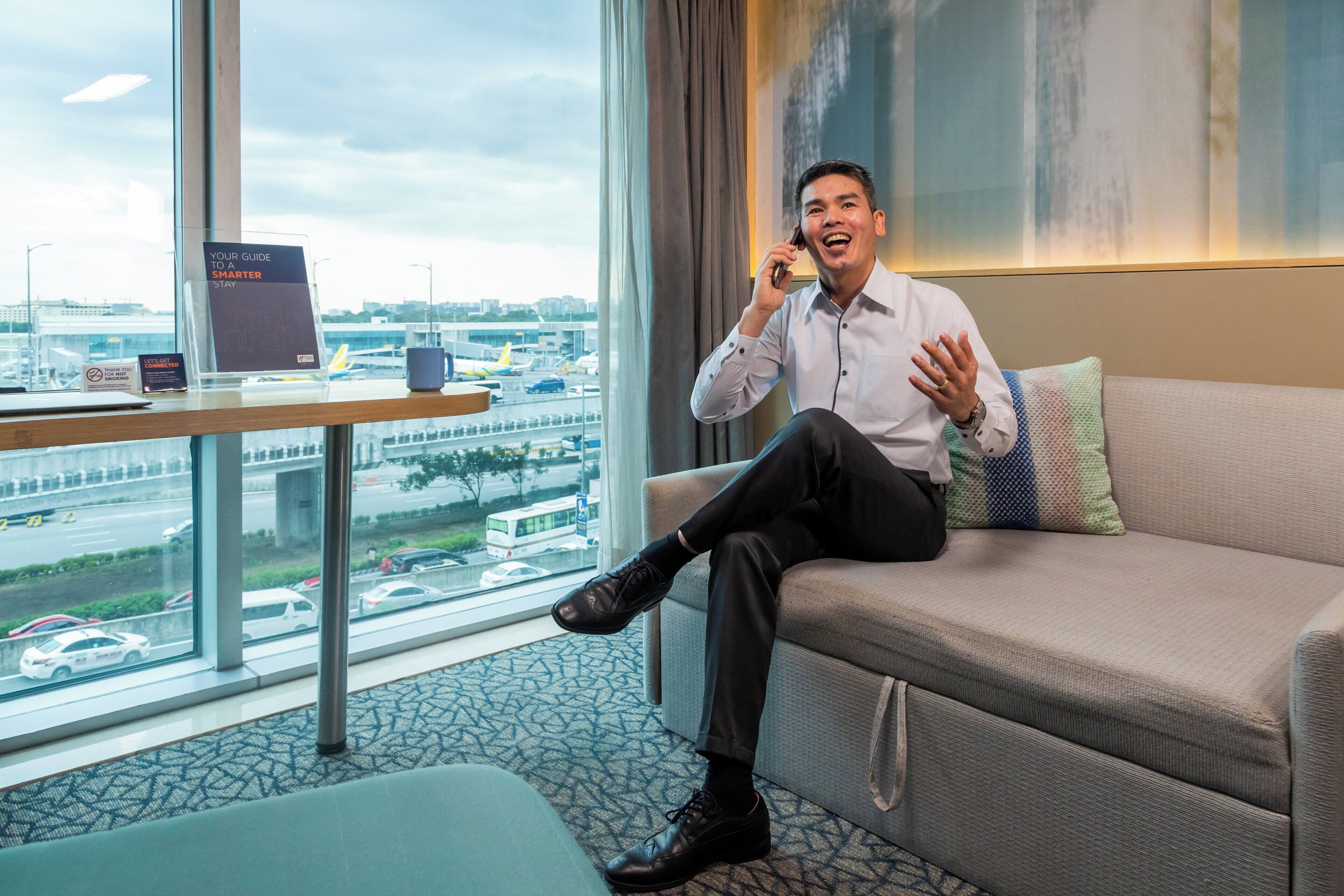 A man sitting on a sofa in a hotel room with a city view, a table with a sign, and a window.