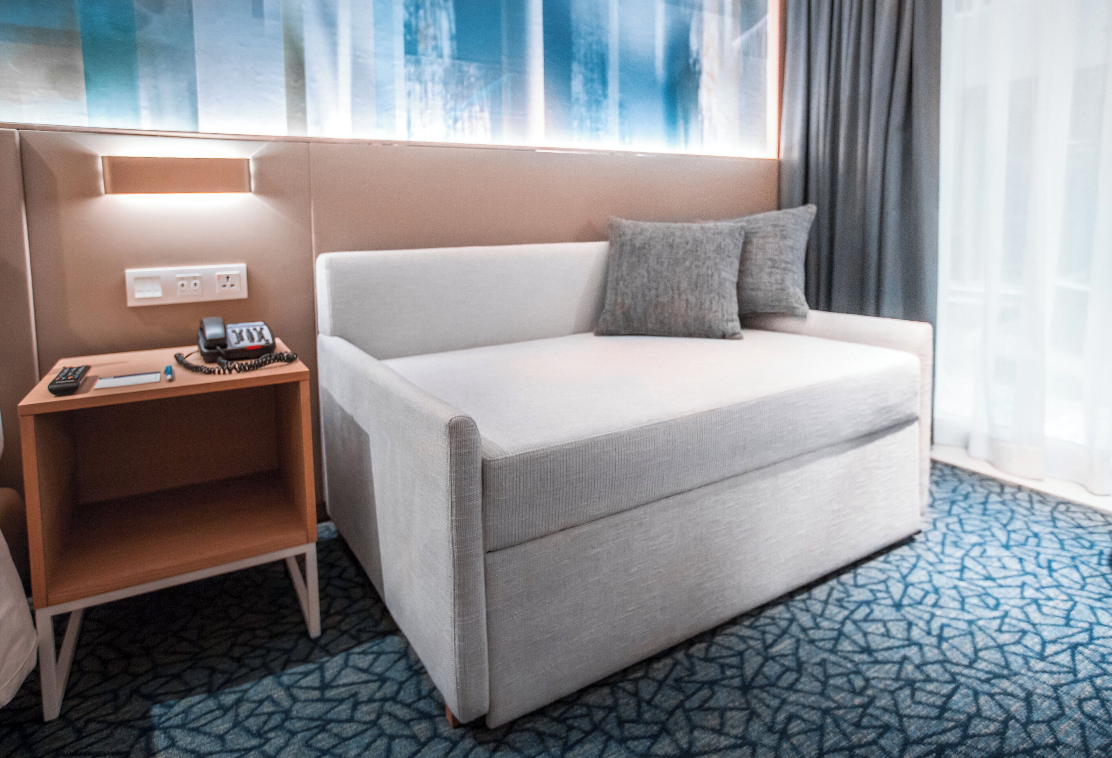 A hotel room with a white sofa, a wooden nightstand with a phone and papers, and a patterned carpet.