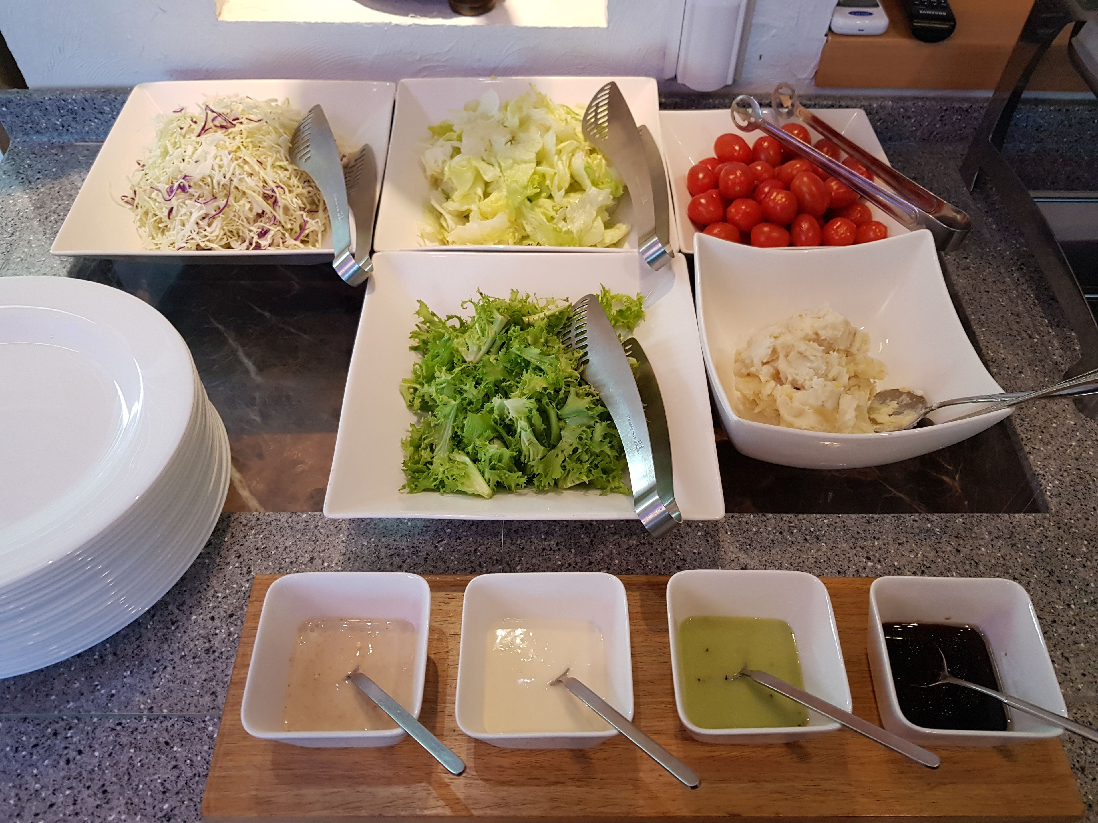 Una mesa con diversos alimentos y condimentos, como ensalada de col, repollo rallado, lechuga, tomates cherry y recipientes con diferentes salsas.