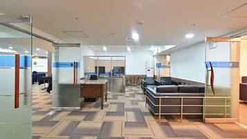 A modern hotel lobby with glass partitions, a reception desk, a waiting area with a sofa, and a patterned carpet.