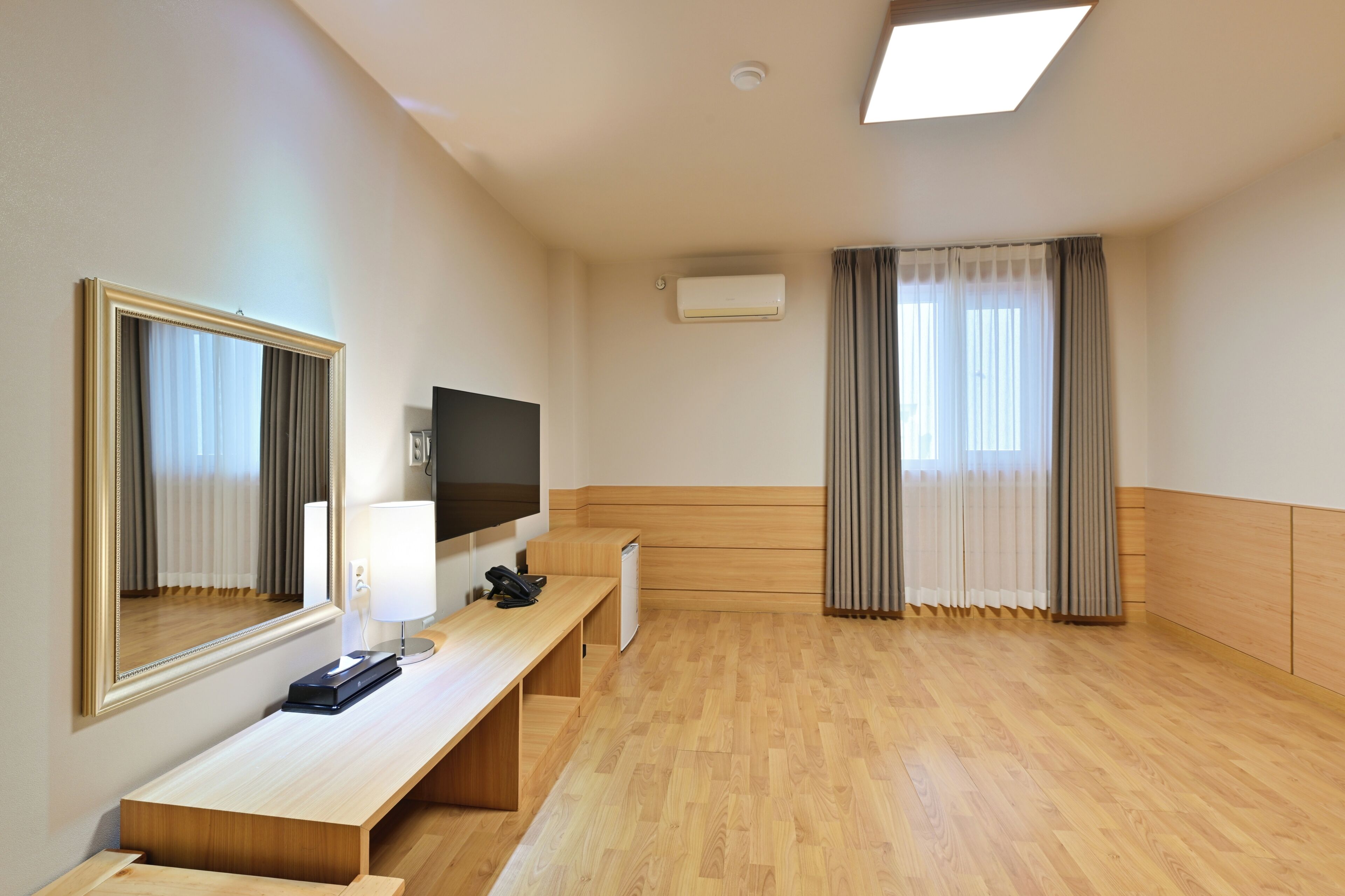 Habitación de hotel con piso de madera, un escritorio con lámpara, televisor, espejo y ventana con cortinas.