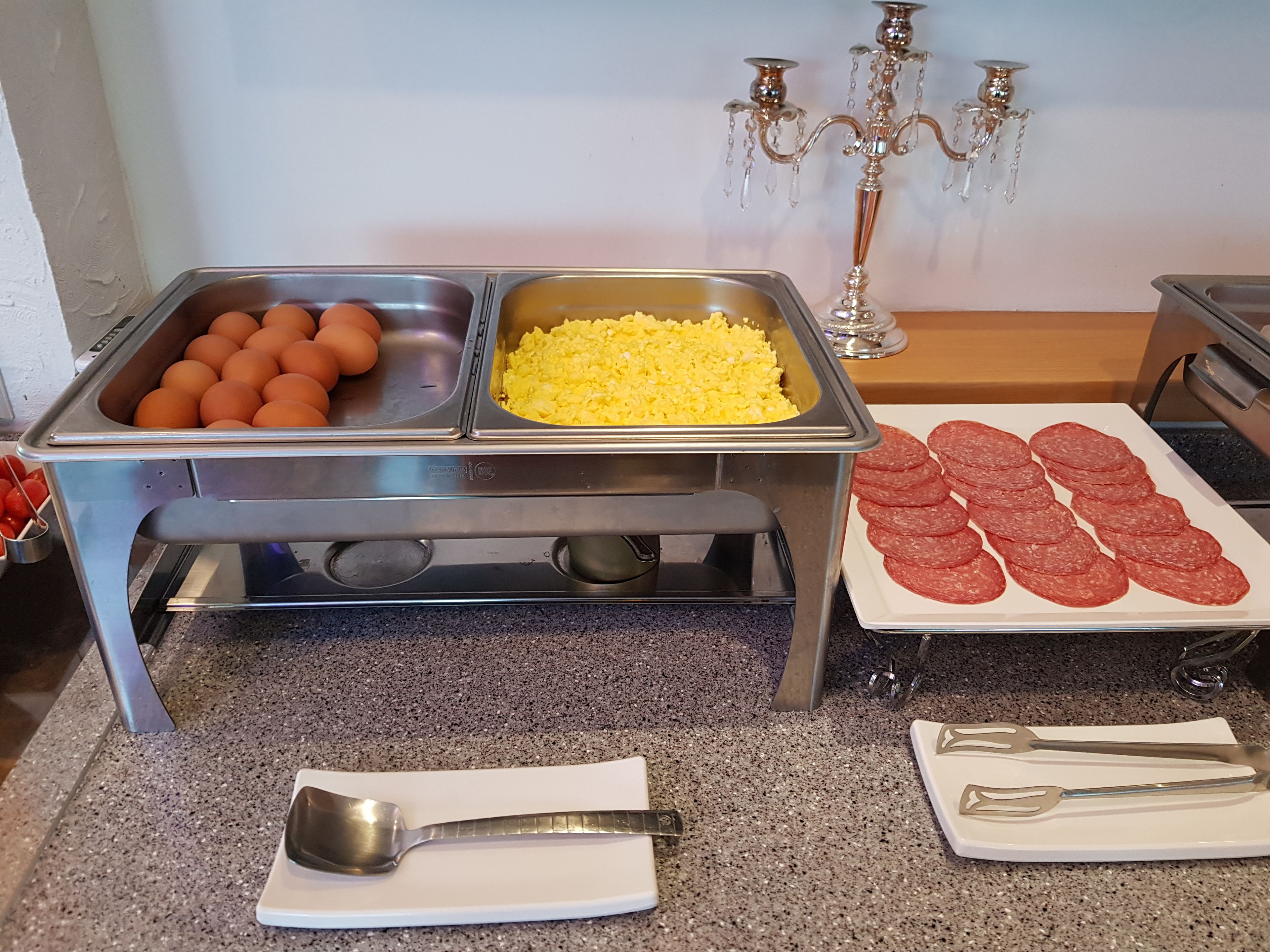 A buffet setup with eggs, scrambled eggs, and sliced salami.