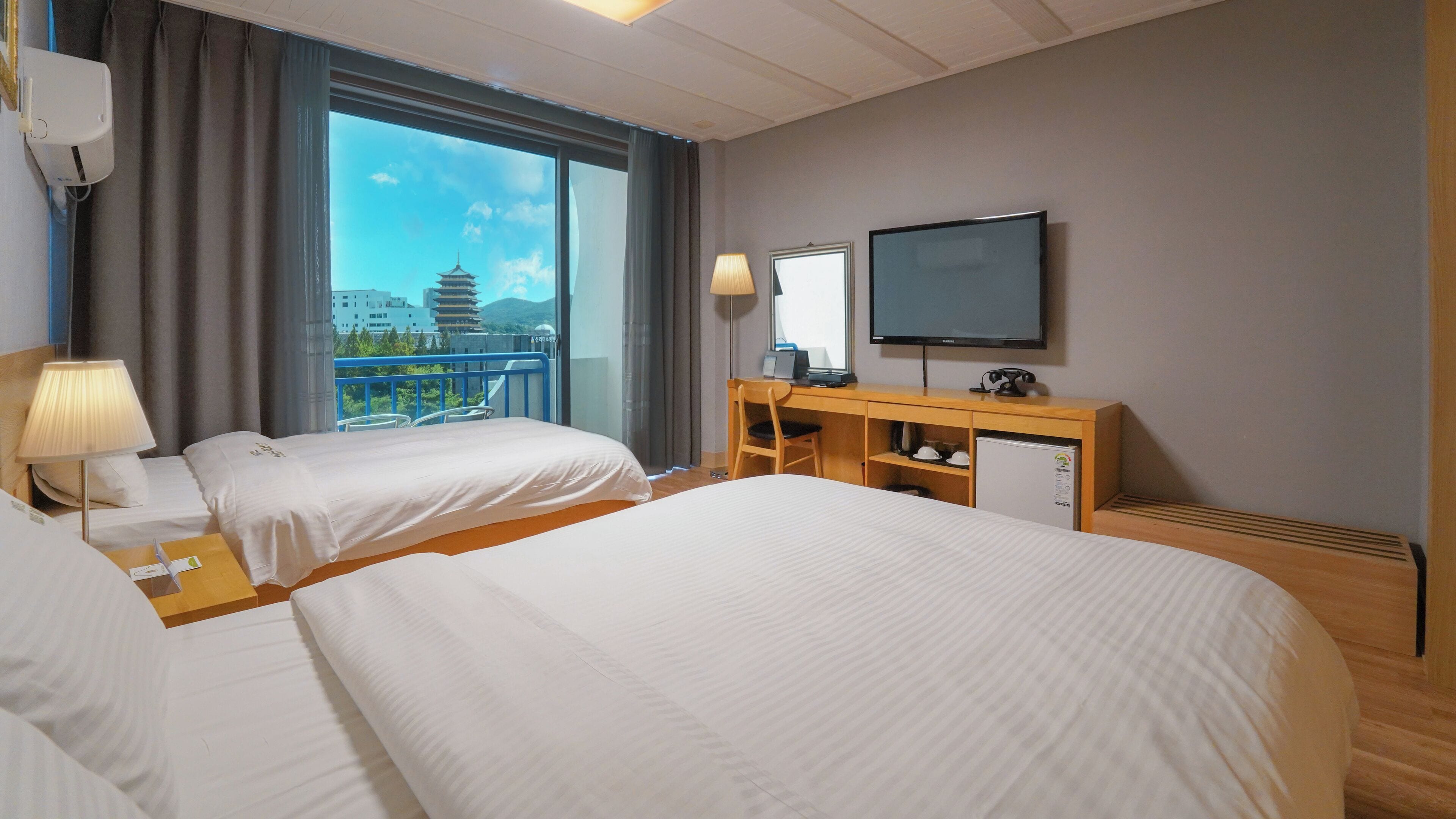 A hotel room with two beds, a desk with a computer, a television, and a view of a pagoda through large windows.