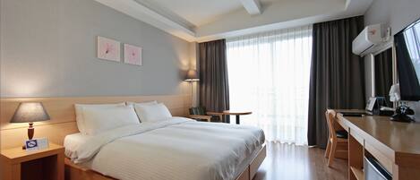 A hotel room with a large bed, a desk, a chair, a lamp, and an air conditioning unit.
