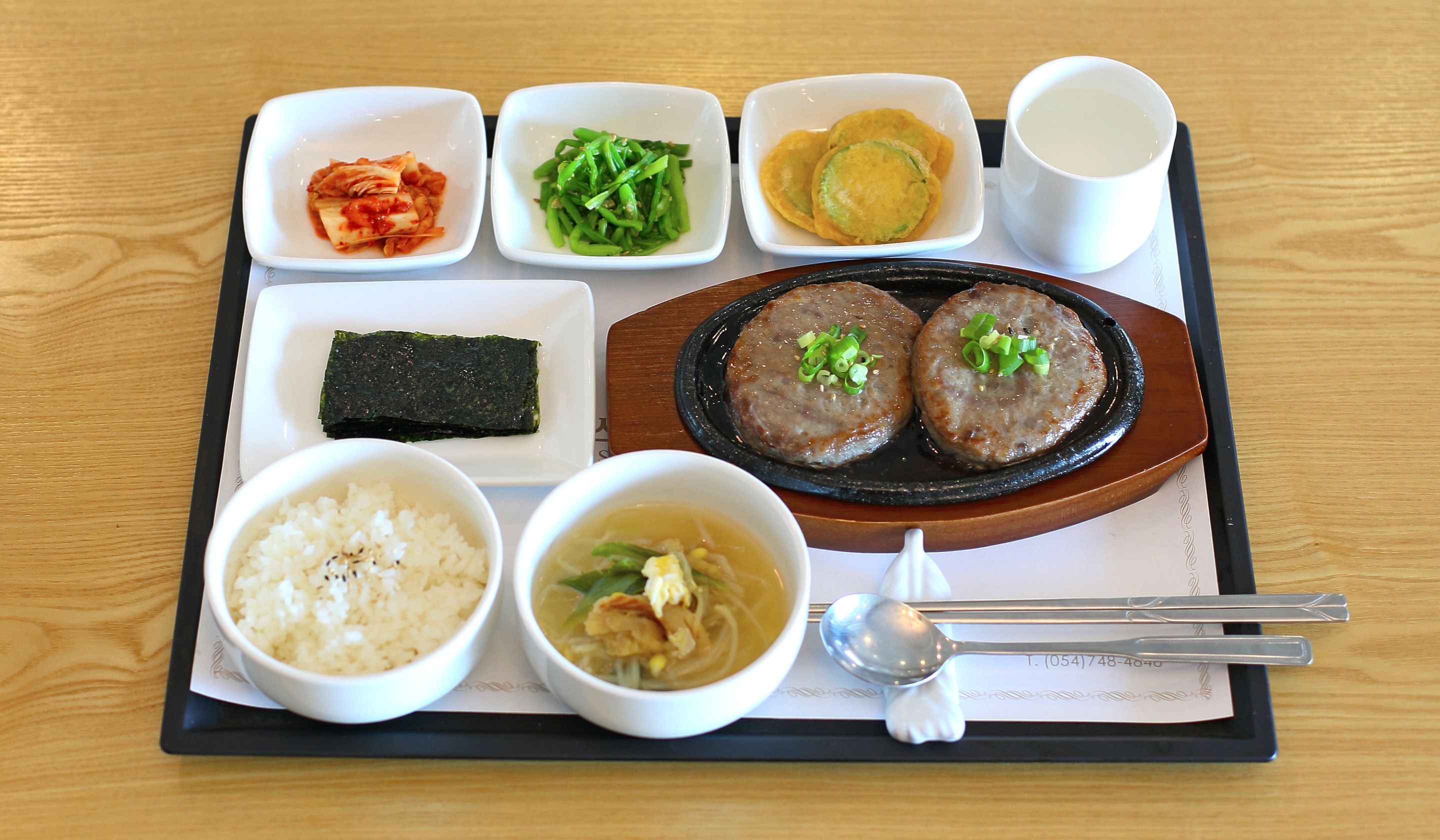A tray with various dishes including rice, soup, kimchi, green vegetables, fried items, and two patties, served on a wooden table.