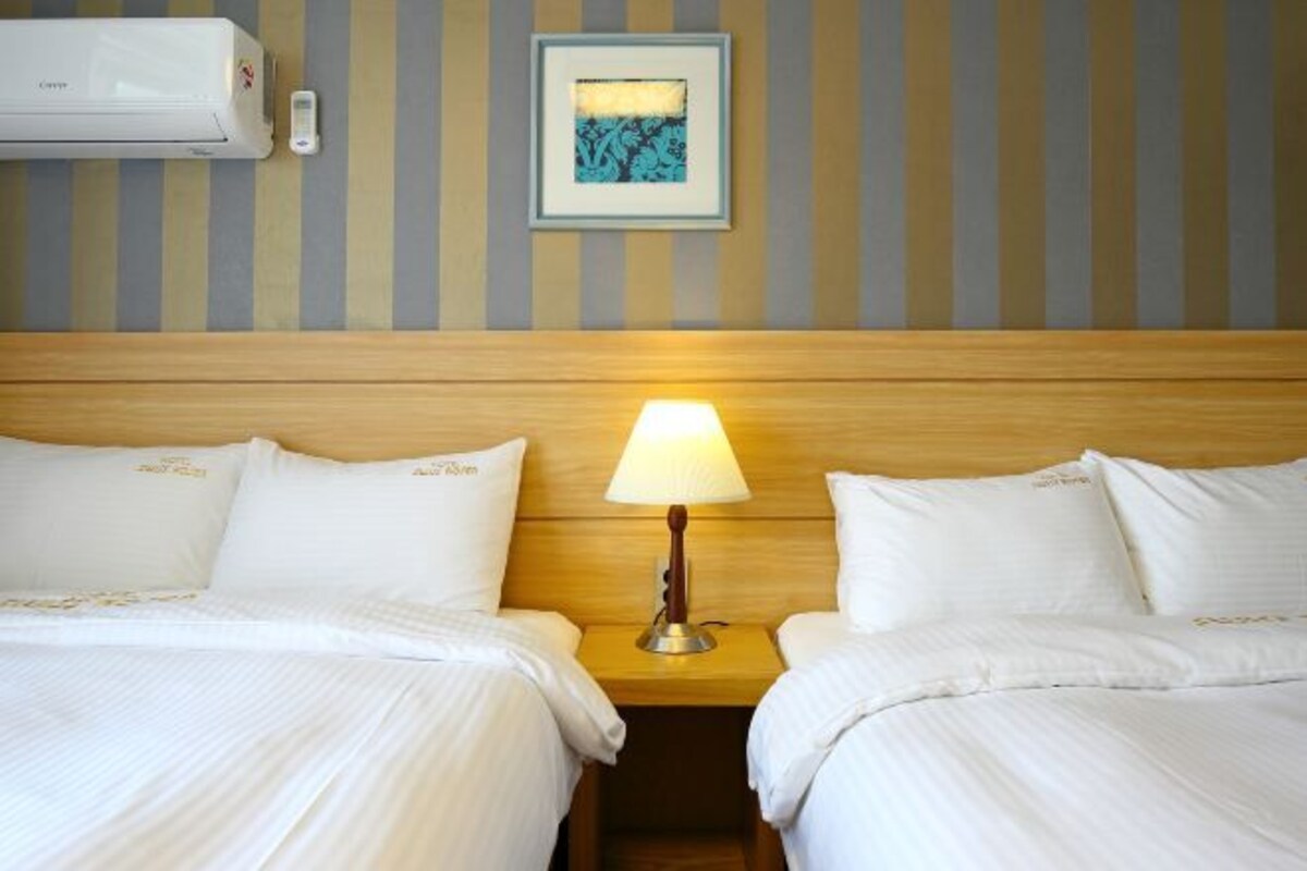 Quarto de hotel com duas camas, cabeceira de madeira, ar-condicionado fixado na parede e quadro emoldurado acima da cabeceira.