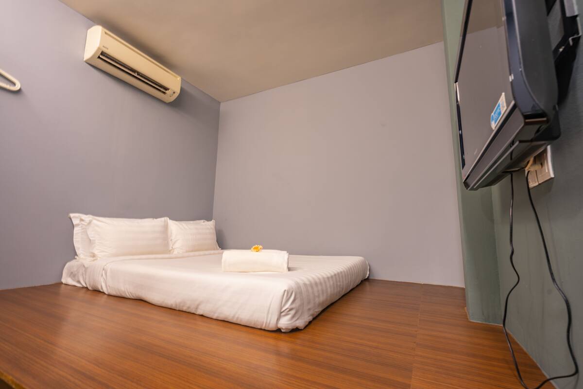 A neatly made bed with white linens, a small bedside table with a flower, and a wall-mounted air conditioner.