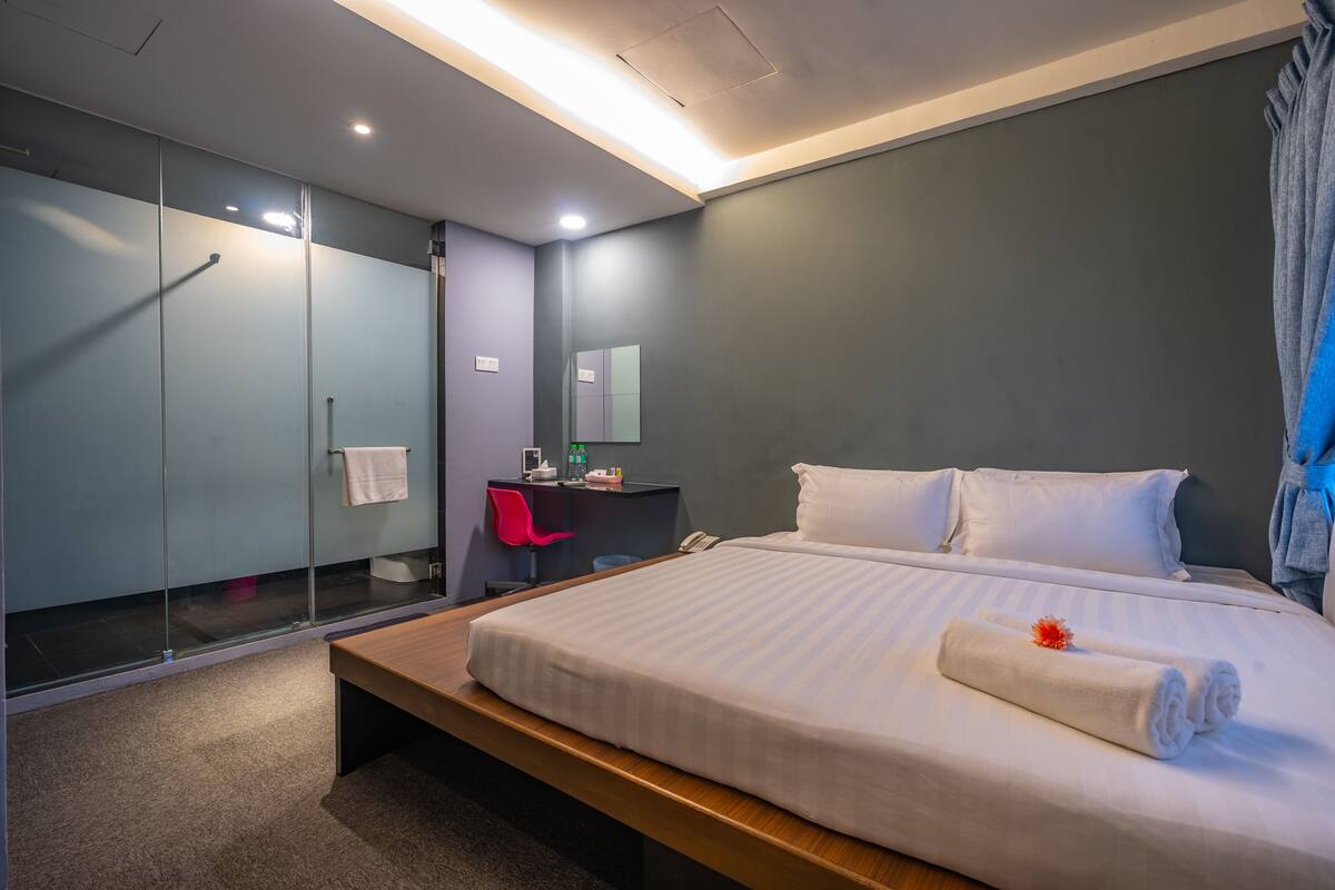 A modern hotel room with a large bed, a bedside table, a desk with a chair, a mirror, and a wardrobe.
