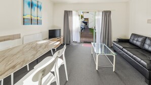 A modern living room with a glass coffee table, a black leather sofa, a wooden dining table with chairs, and a flat-screen TV. - Birchwood Spa Motel (Rotorua)