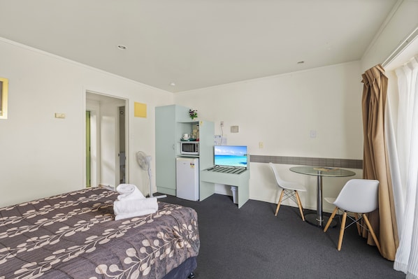 A hotel room with a bed, a desk with a computer, a chair, a microwave, and a refrigerator.