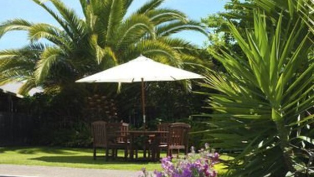 A patio area with a table and chairs under a white umbrella, surrounded by purple flowers and green foliage. - Birchwood Spa Motel (Rotorua)
