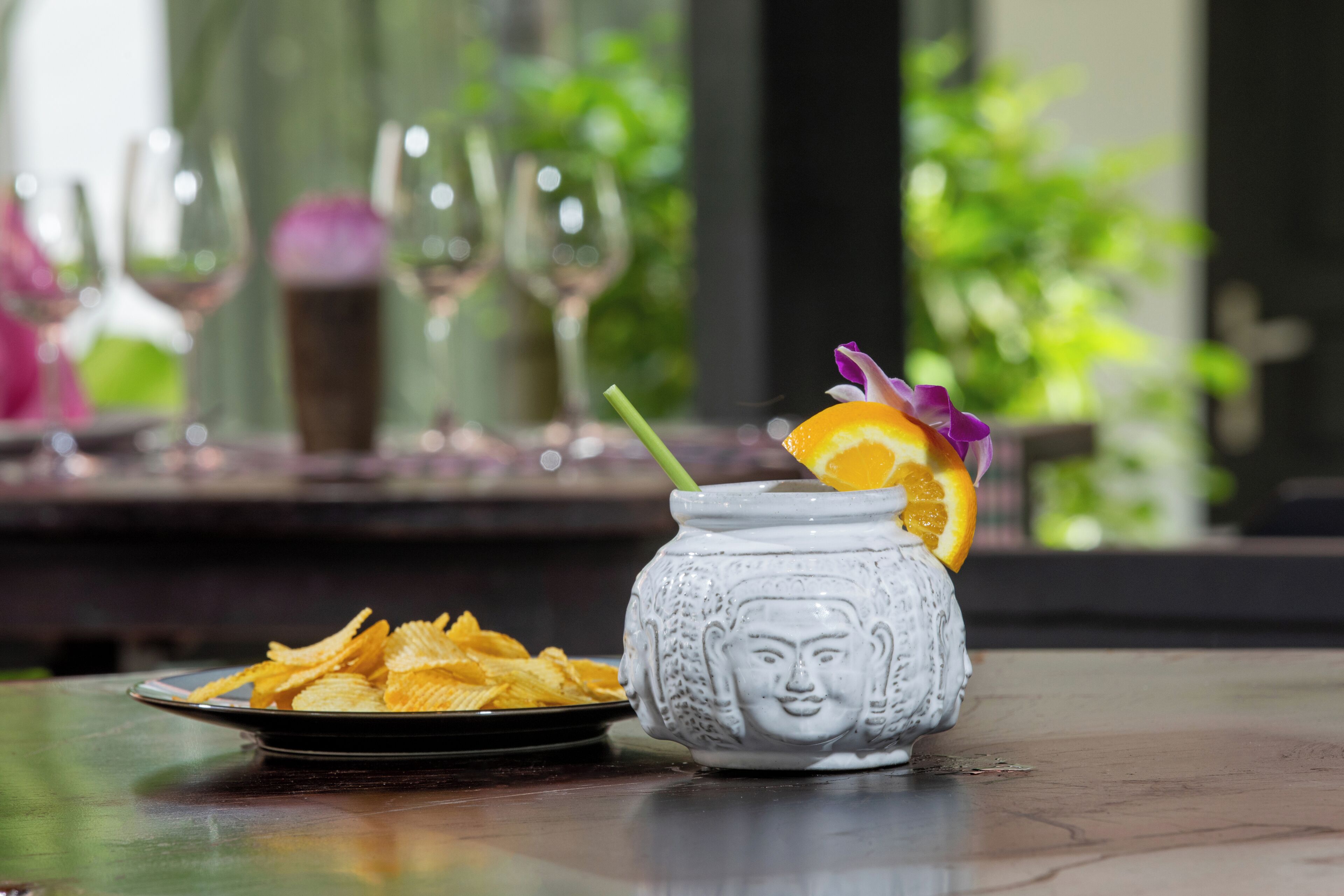 A white ceramic mug with a face design, filled with a beverage and garnished with an orange slice and a purple flower. A plate of potato chips is visible in the foreground.