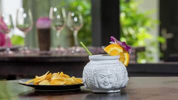 Una taza de cerámica blanca con un diseño facial, llena de una bebida y adornada con una rodaja de naranja y una flor morada. En primer plano, se ve un plato de papas fritas.