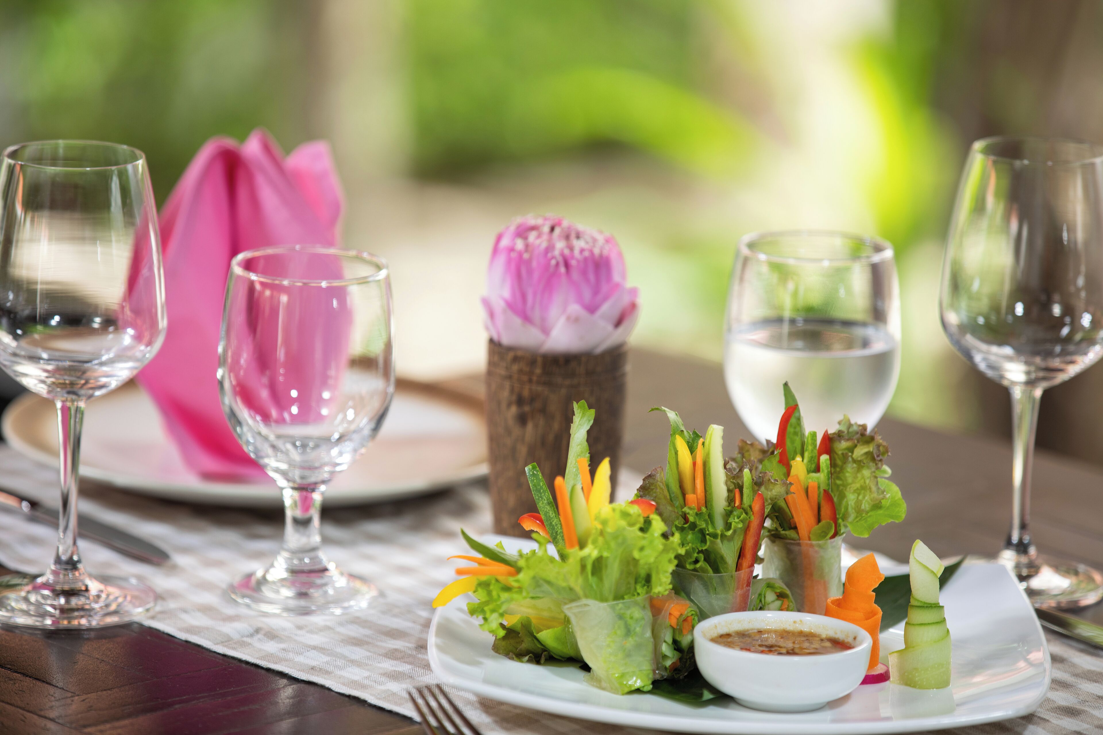 A table set with clear glasses, a pink napkin, a floral centerpiece, and a plate with a vegetable salad and dipping sauce.