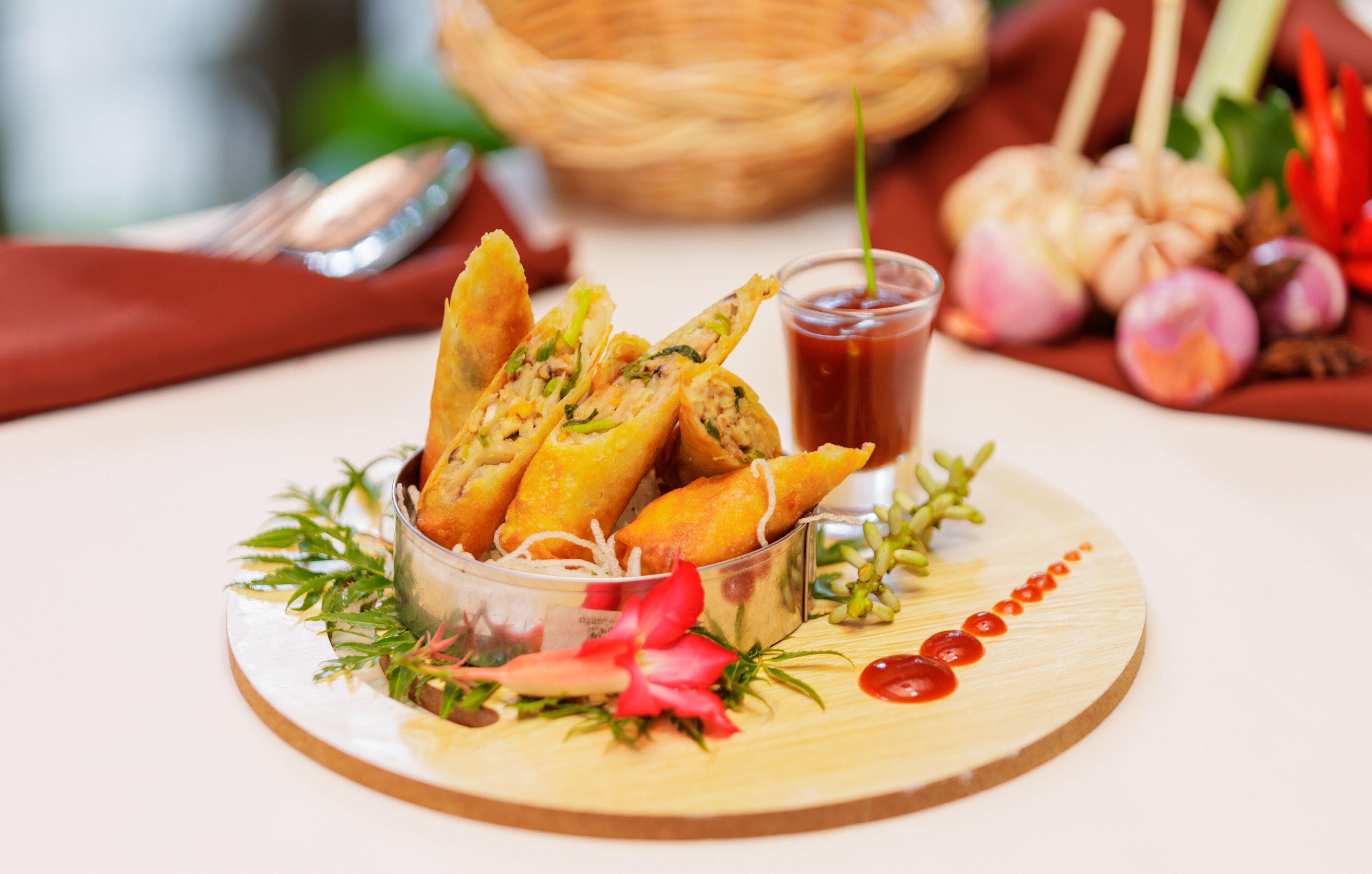 A wooden board with a clear glass bowl containing fried spring rolls, garnished with herbs and a small dish of sauce.
