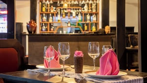 Un espace bar avec des étagères remplies de bouteilles et de verres, un comptoir et une table dressée avec des verres à vin et un vase orné d’une fleur rose.