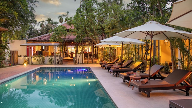 Une piscine entourée de chaises longues et de parasols, elle-même bordée d’une végétation luxuriante et d’un bâtiment doté d’un toit en tuiles.