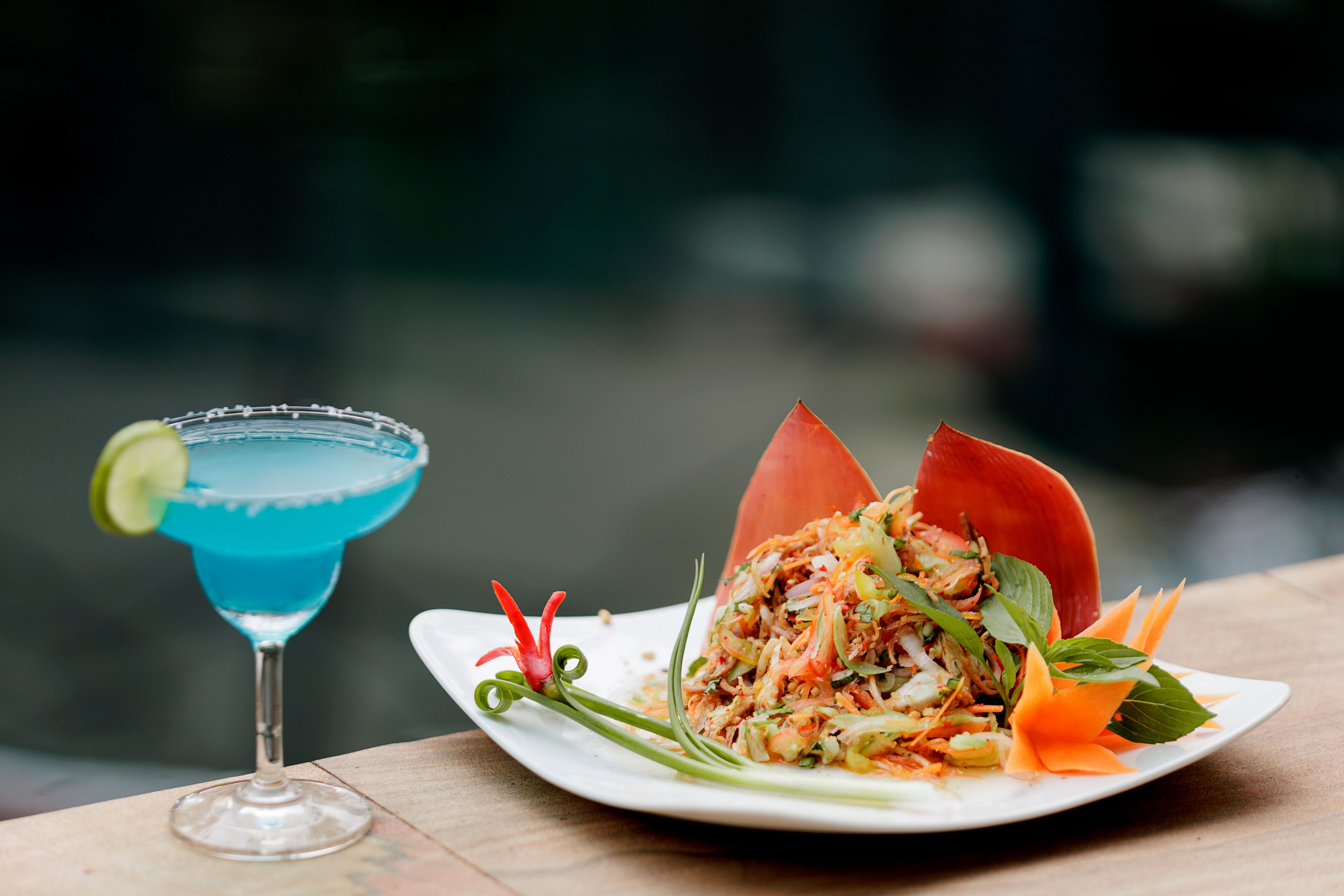 A blue cocktail with a lime garnish and a plate of salad with vegetables and herbs.