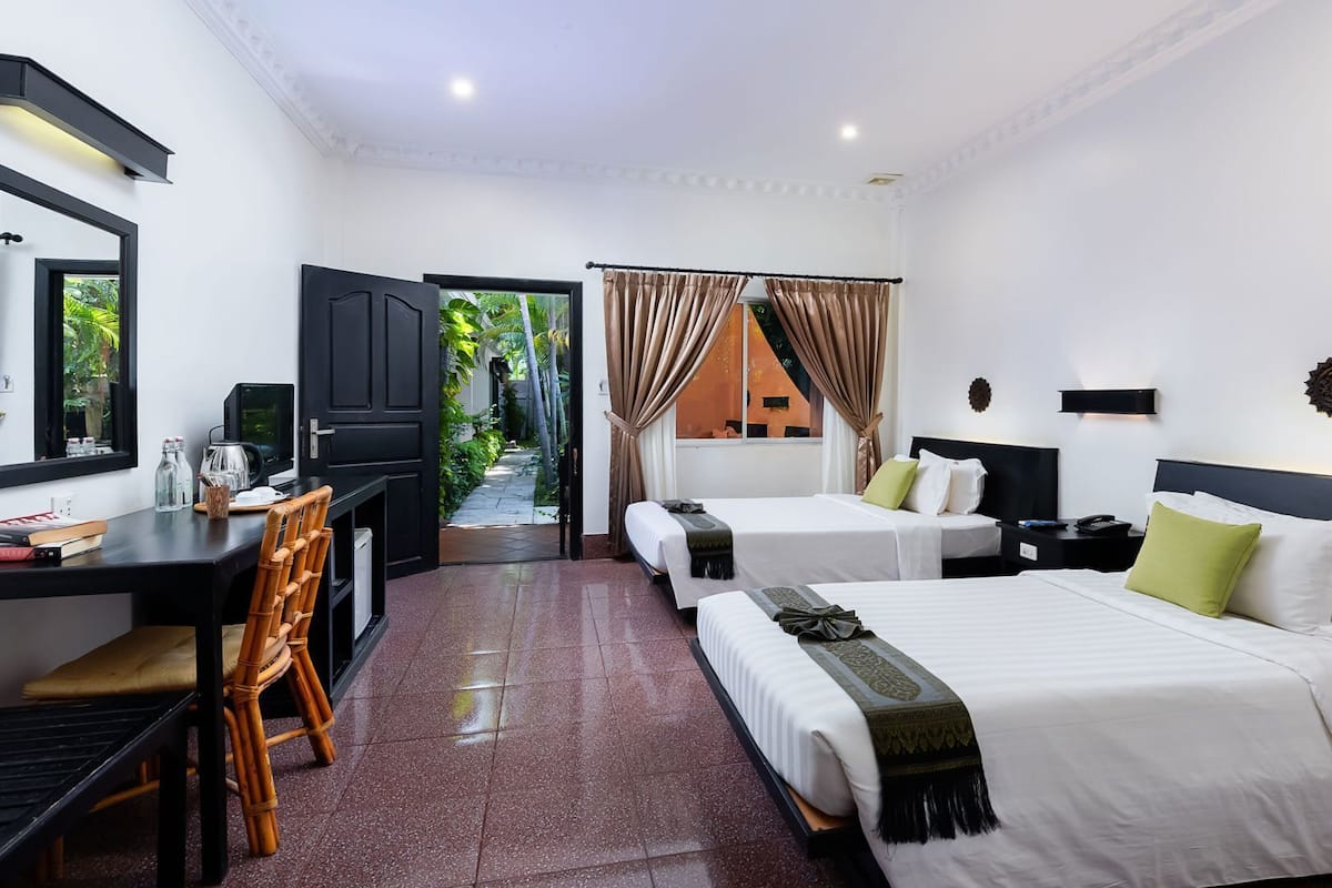 A hotel room with a desk, two beds, a view of a garden, and a mirror.