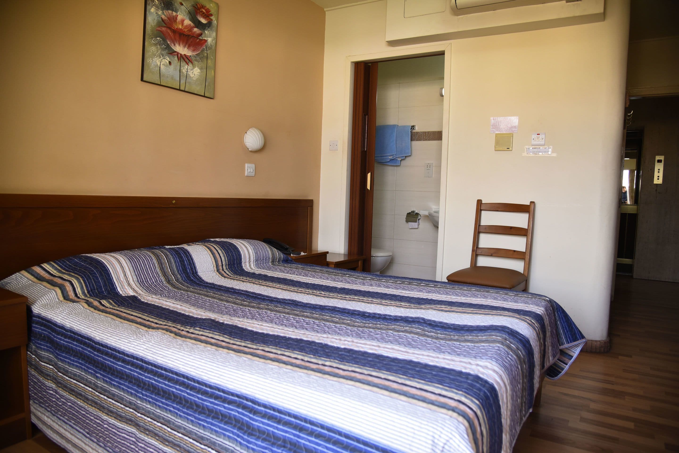 A bedroom with a large bed, a wooden headboard, a nightstand, a chair, and a bathroom visible through an open door.