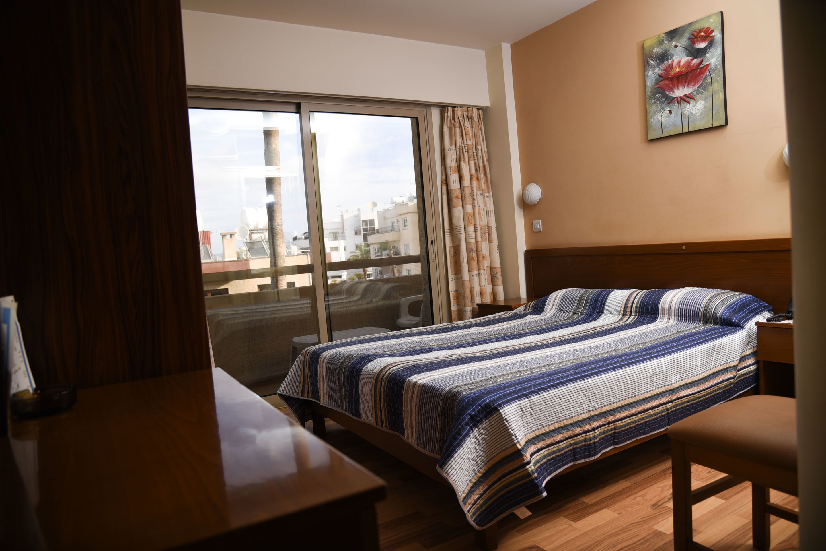 A hotel room with a bed, a desk, a chair, a window with curtains, and a painting on the wall.