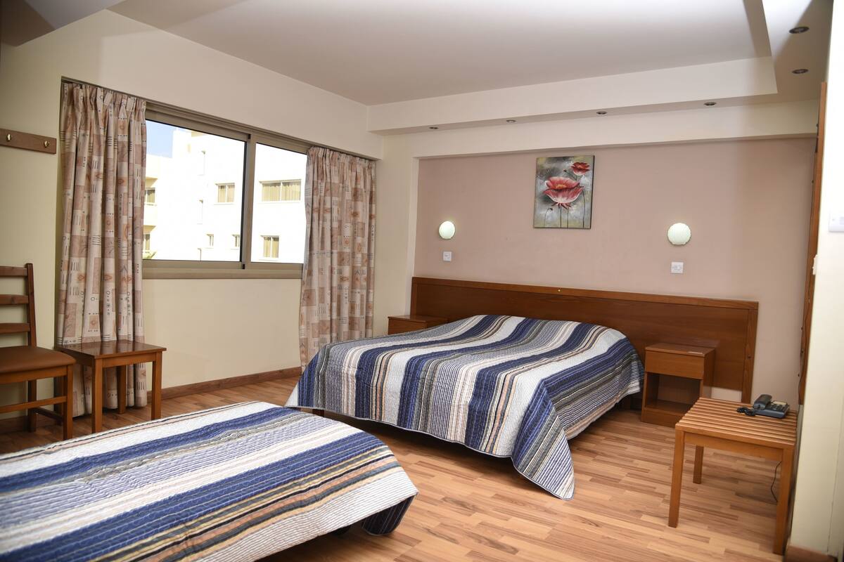 Une chambre d’hôtel avec deux lits, des meubles en bois, une fenêtre avec des rideaux, un tableau au mur et des lampes de chevet.