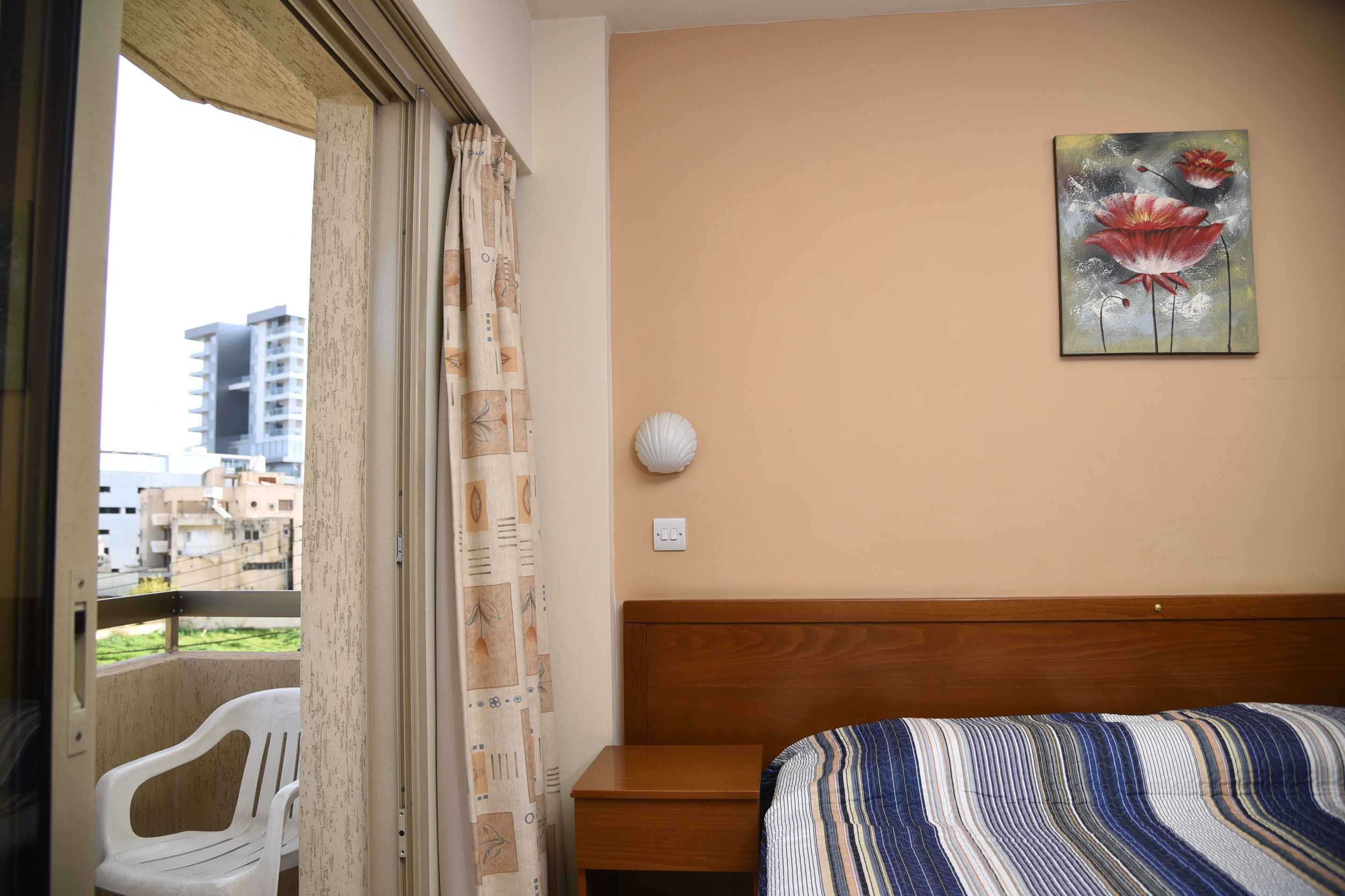 A hotel room with a bed, a nightstand, a wall-mounted lamp, a window with a view of buildings, and a curtain.