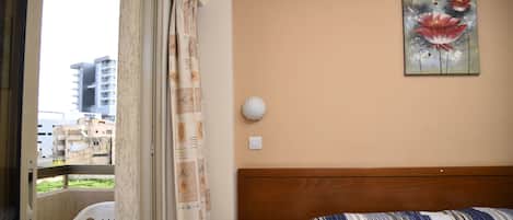 A hotel room with a bed, a nightstand, a wall-mounted lamp, a window with a view of buildings, and a curtain.