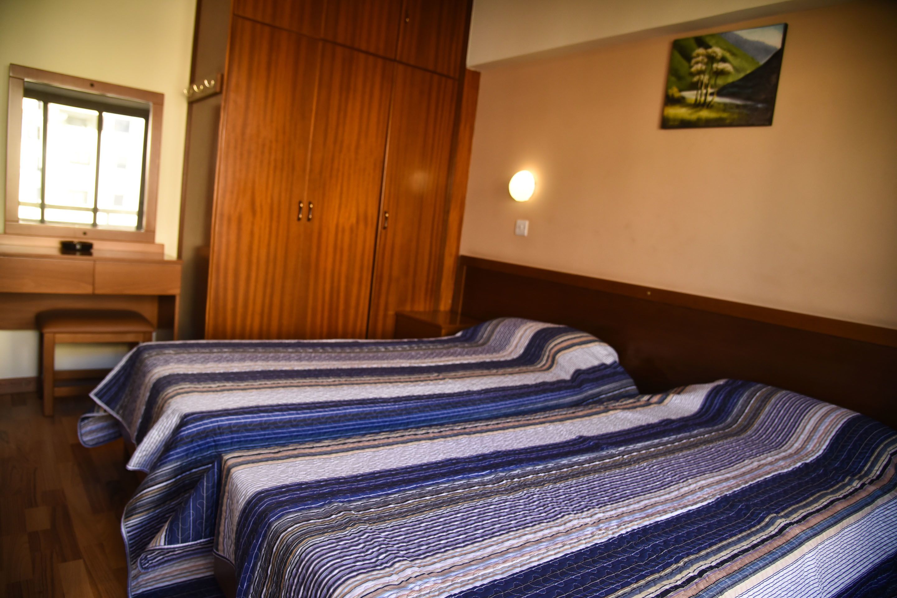 A bedroom with two beds, a wooden wardrobe, a desk, and a window.