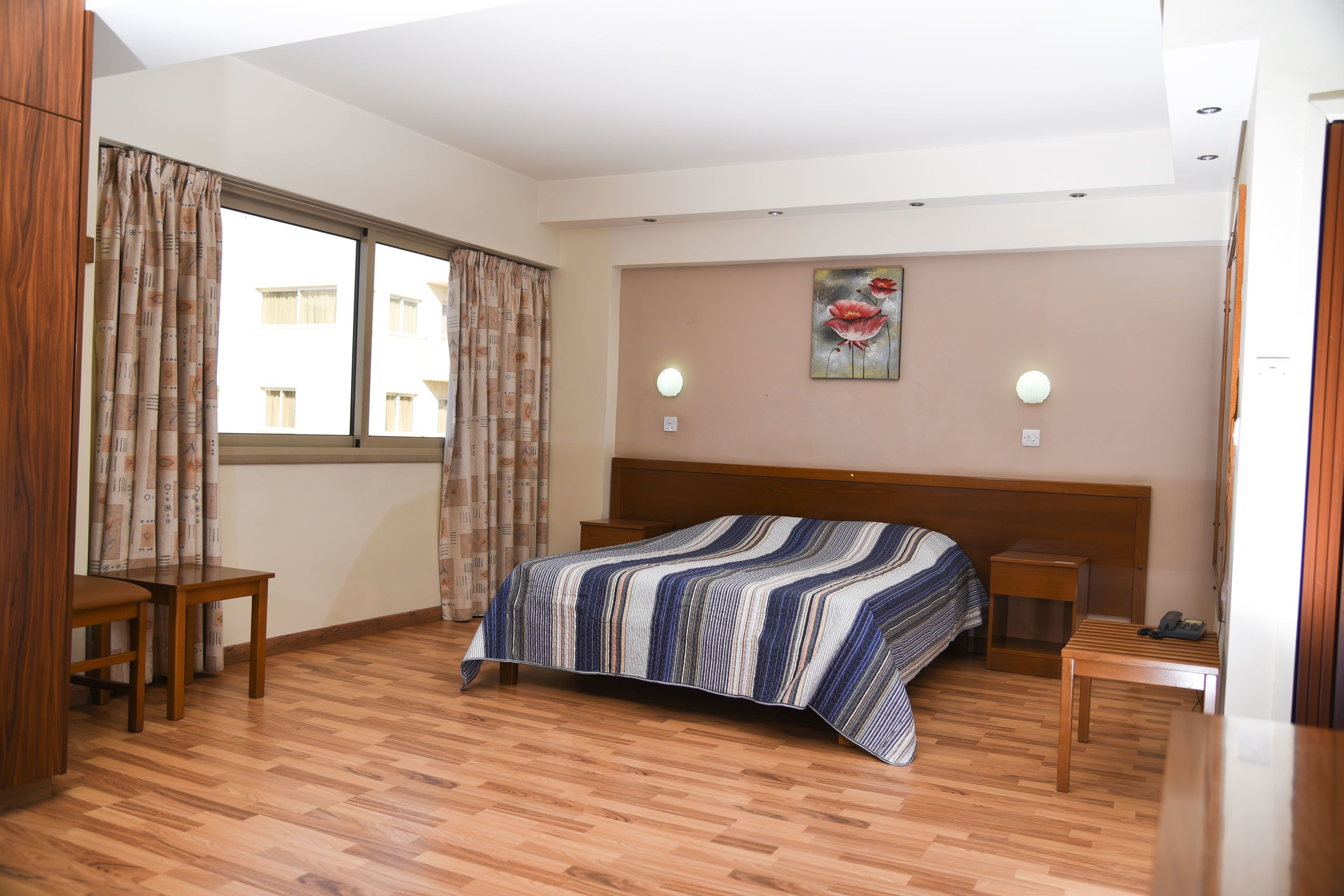 Une chambre d’hôtel avec un plancher en bois, un lit recouvert d’une couvre-lit à rayures, une table de chevet en bois, un bureau et une chaise. Il y a deux fenêtres avec des rideaux, un luminaire fixé au mur et un tableau accroché au mur.