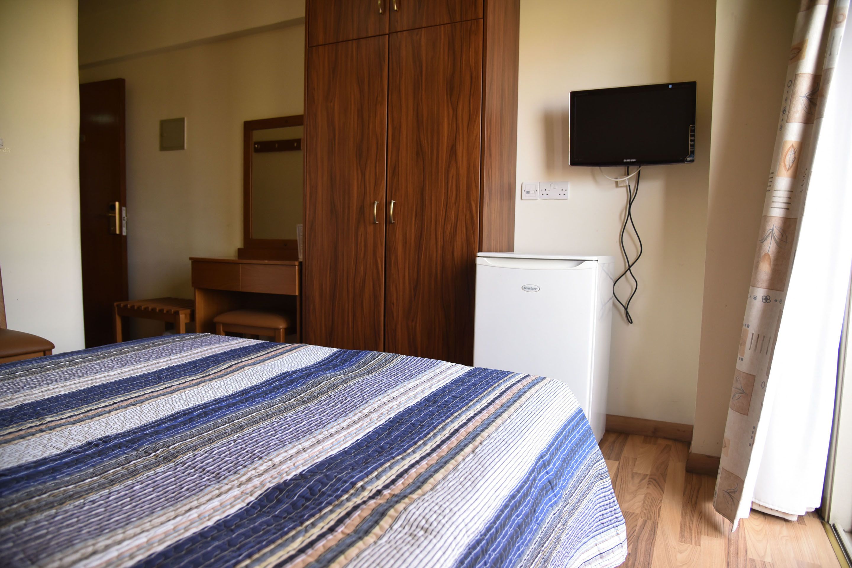 A bedroom with a bed, a wooden wardrobe, a small white refrigerator, and a mounted television.