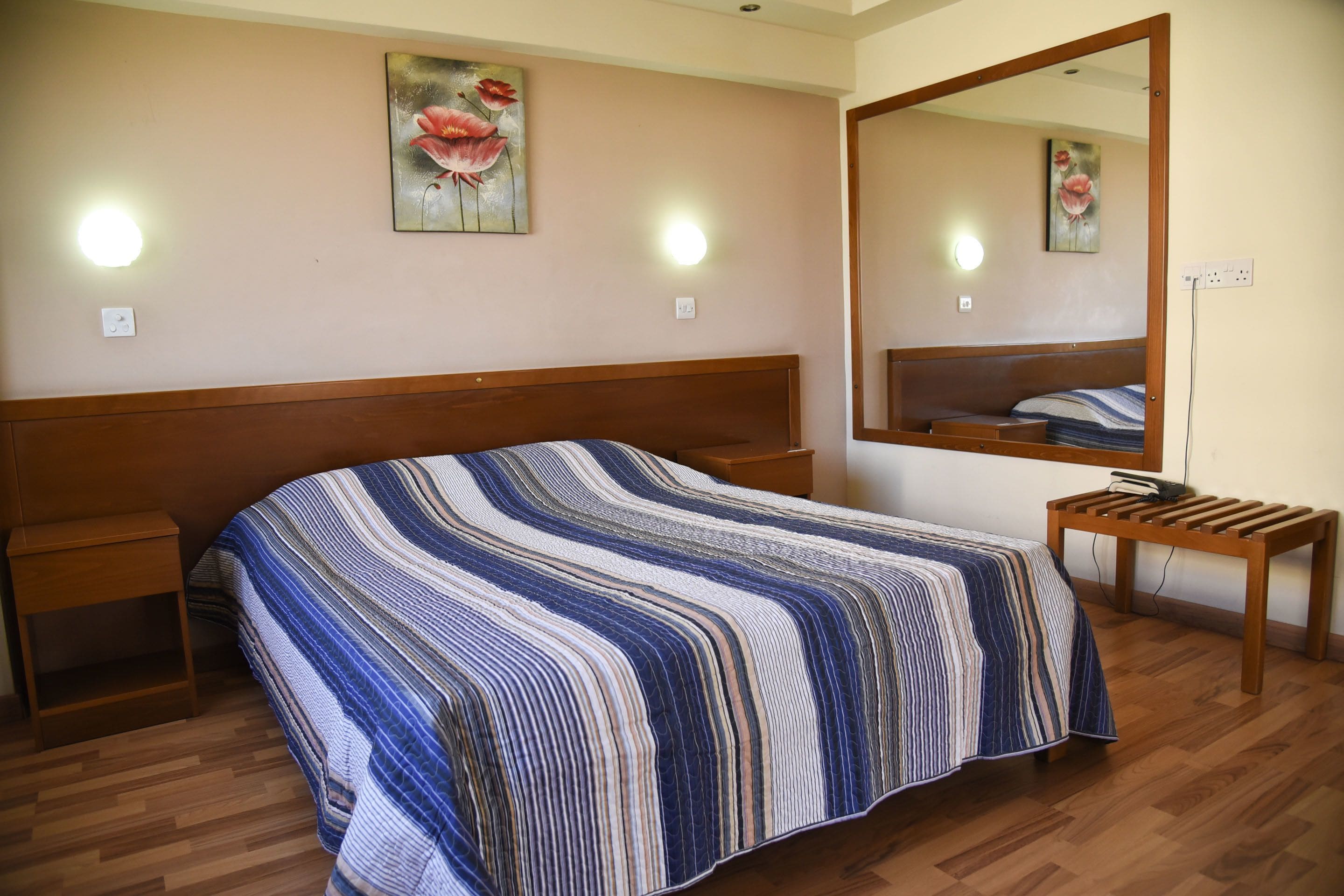 Un dormitorio con una cama de madera, una mesita de noche, un banco, un espejo grande y dos luces fijadas a la pared.