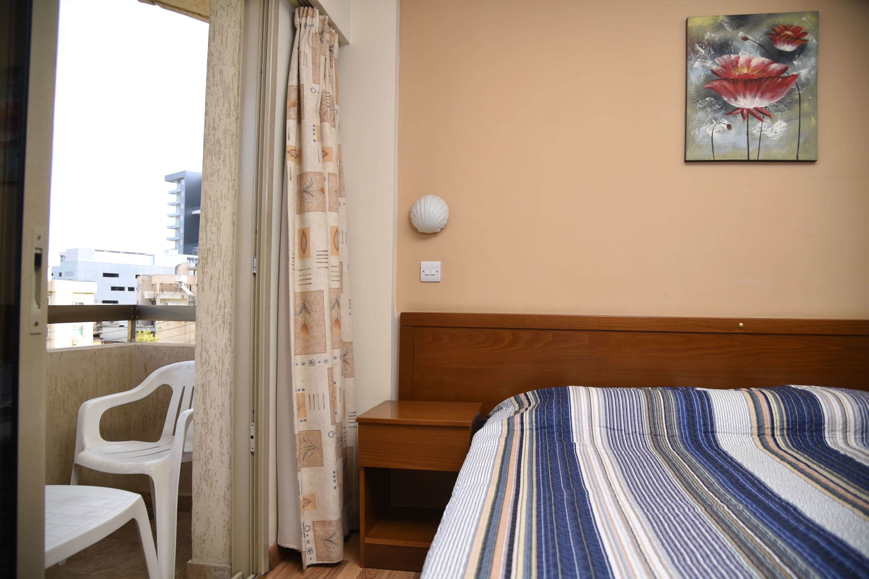 Habitación de hotel con una cama de madera, una colcha a rayas, una silla de plástico blanco, una mesita de noche de madera, una lámpara de pared y vistas a edificios a través de una ventana con cortinas.