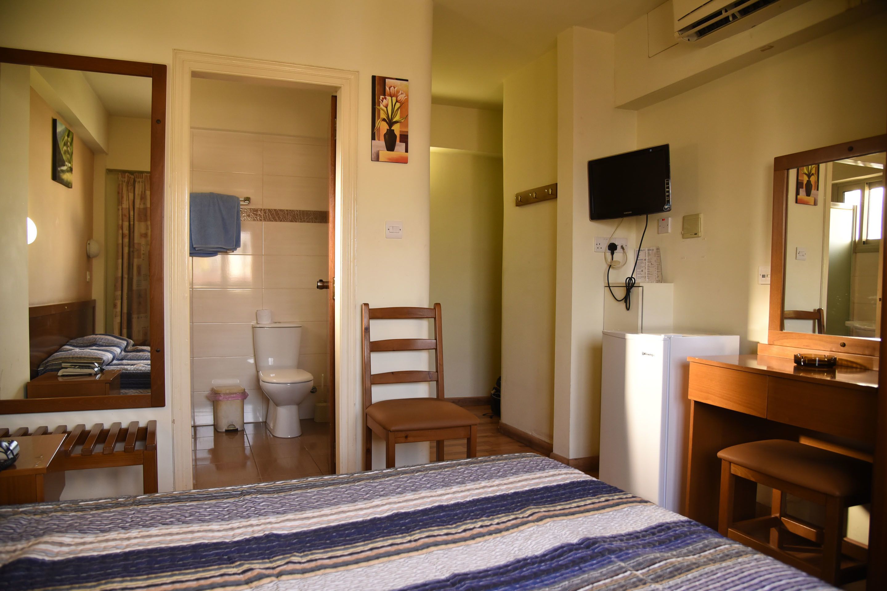 Habitació d'hotel amb un llit, una cadira de fusta, escriptori, televisió, mirall, bany amb vàter i cortina de dutxa, i nevera.