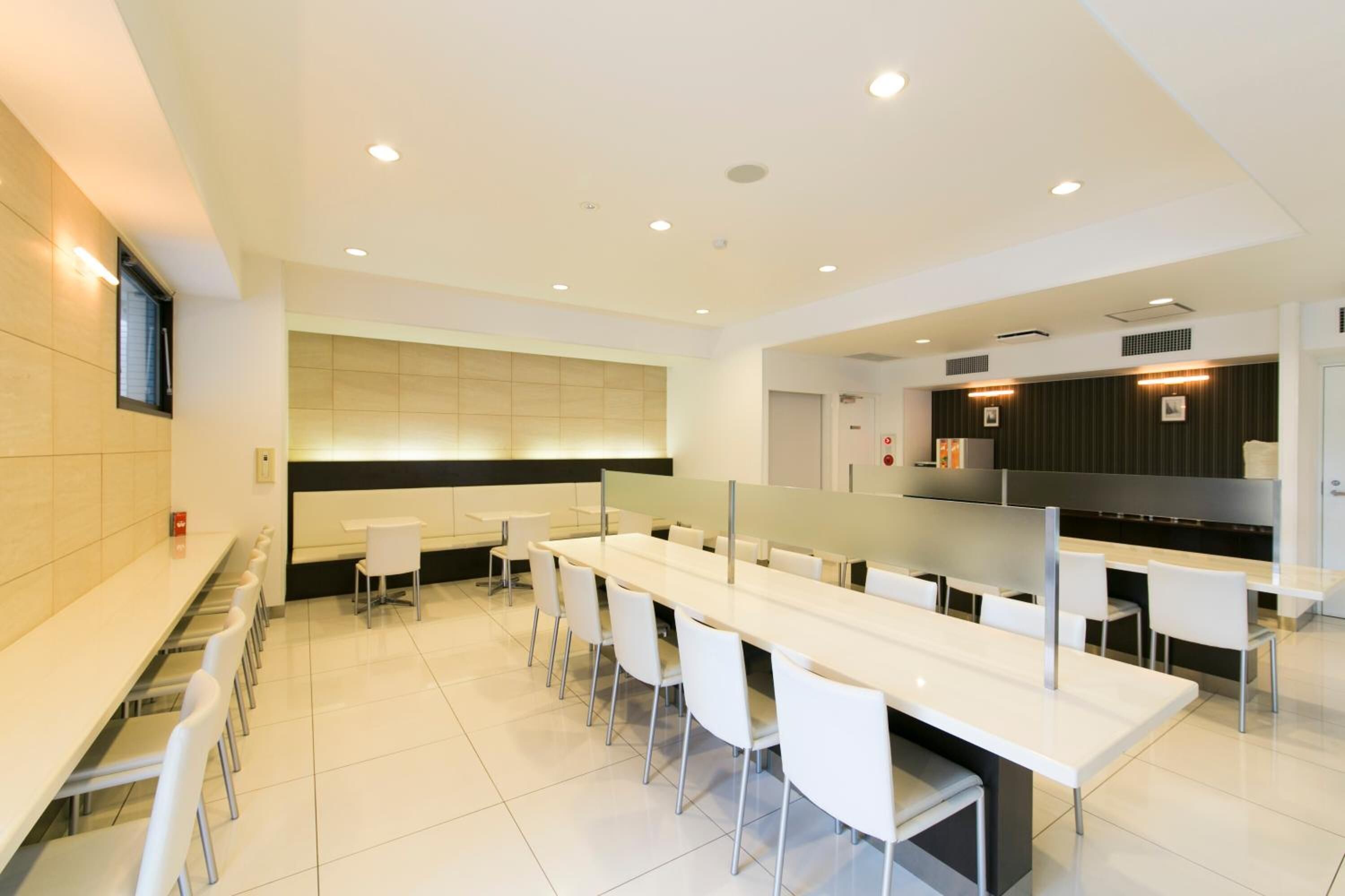 A modern dining area with long white tables and chairs, a counter with a black backsplash, and recessed ceiling lighting.