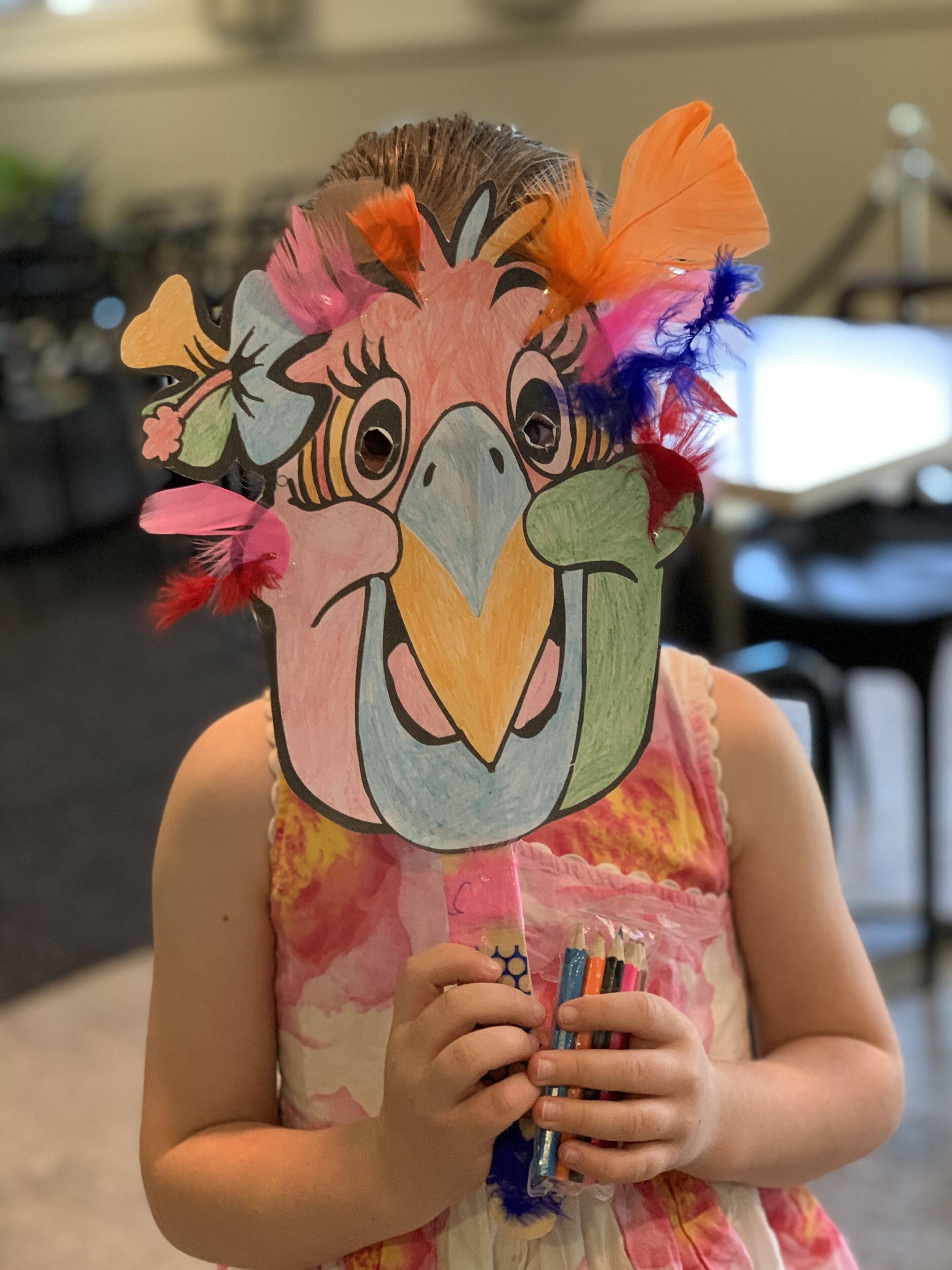 A person holding a colorful mask with feathers and a beak.