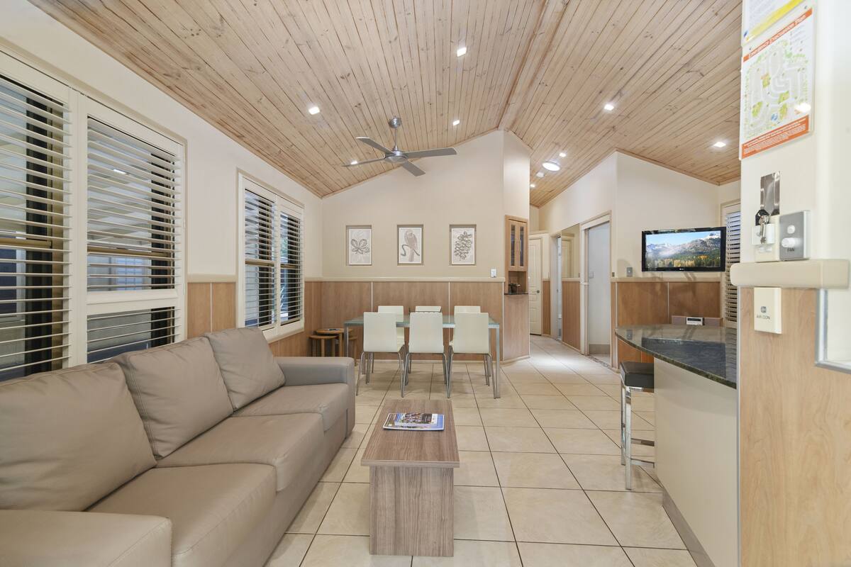 A spacious living area with a wooden ceiling, a beige sofa, a dining table with chairs, and a kitchen area with a TV.