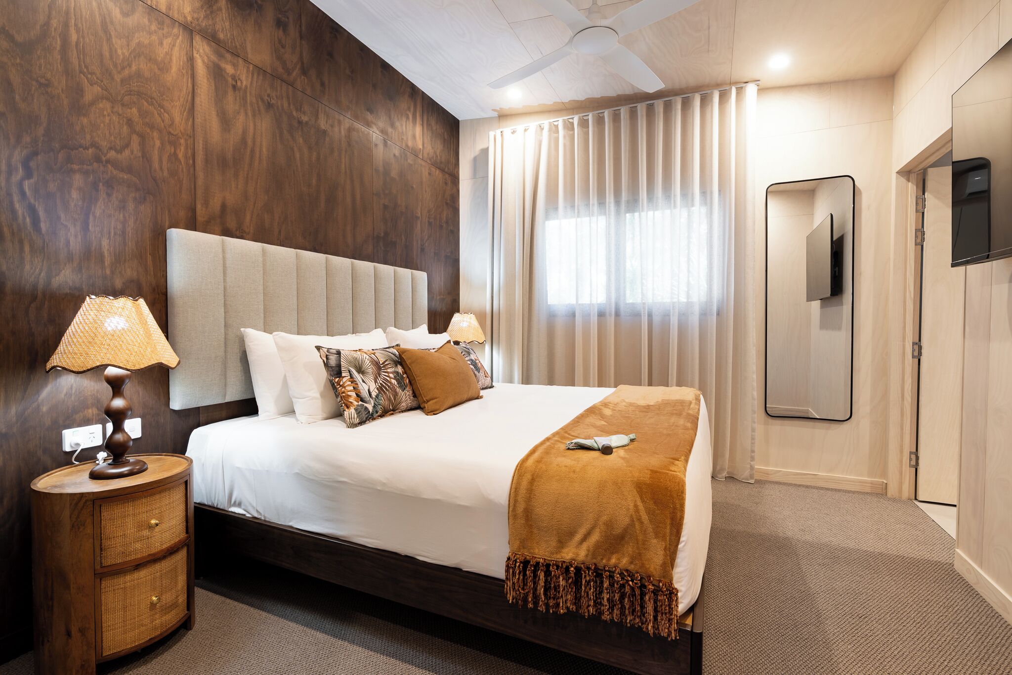 A hotel room with a large bed, a bedside lamp, a wooden nightstand, a mirror, and a window with curtains.