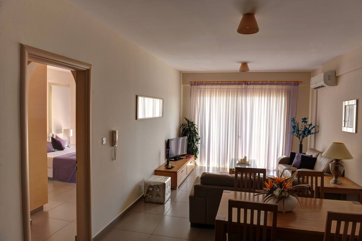 A modern living room with a sofa, TV, and dining table. There is a door leading to a bedroom.