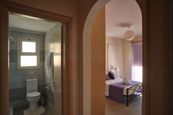 Un baño de hotel con inodoro, ducha y una pequeña ventana. La habitación tiene una cama con una colcha morada y una toalla blanca.