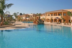 Zona de piscina en un resort con varios edificios, palmeras y una piscina azul cristalina.