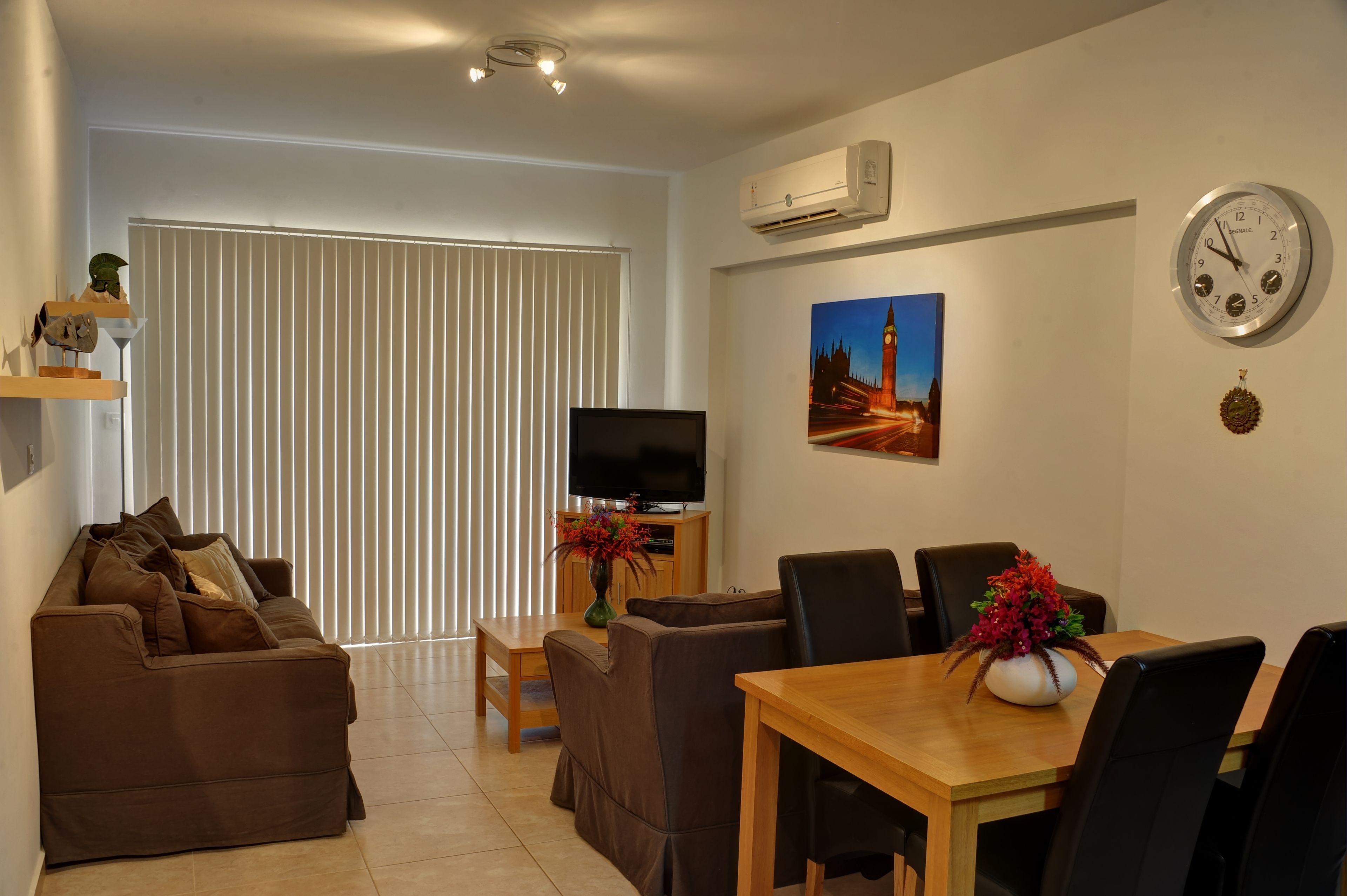 Een woonkamer met een bruine bank, een houten eettafel met stoelen, een televisie op een dressoir, en een schilderij van een stadsgezicht.