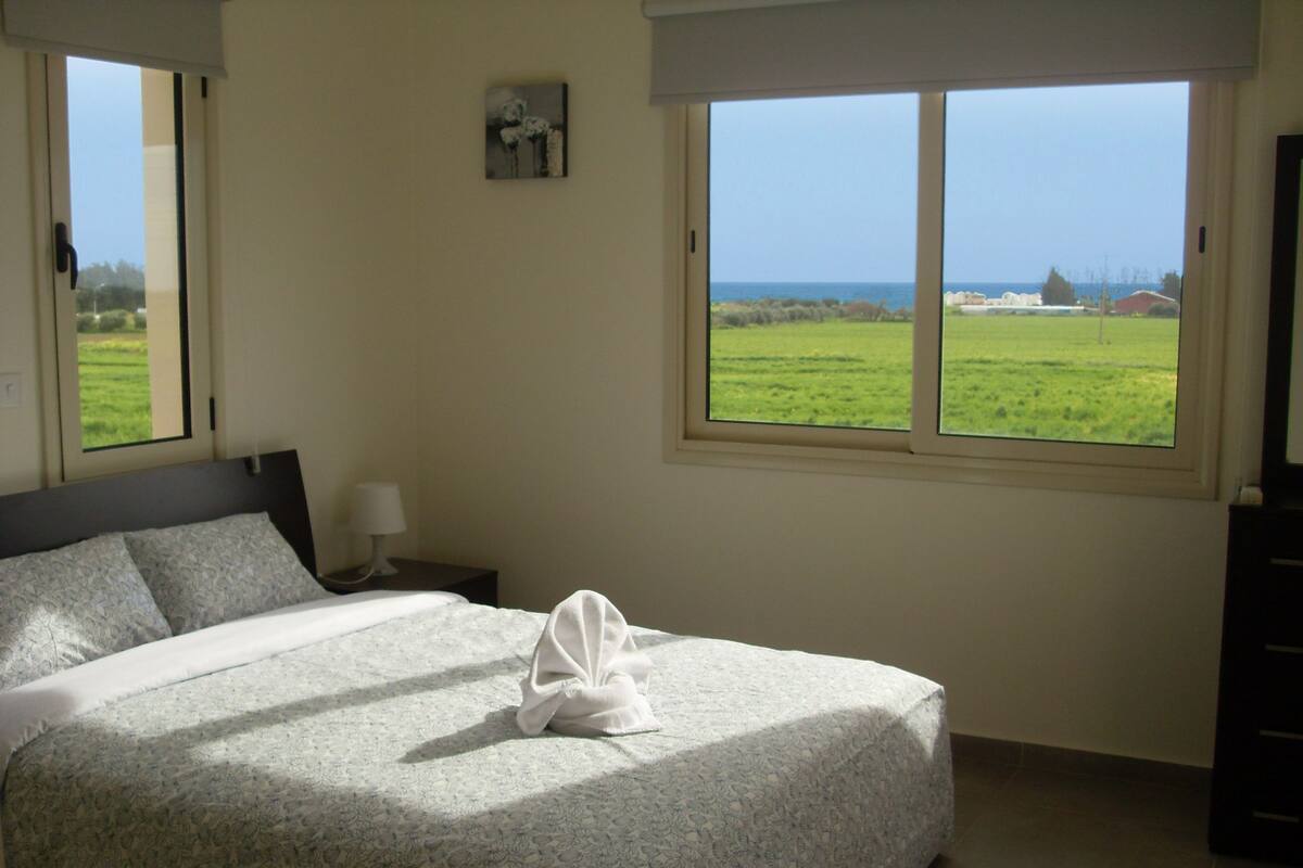 A neatly made bed with a floral bedspread, a bedside lamp, and a view of a green field through a window.