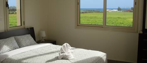 Una cama bien hecha con una colcha estampada con motivos florales, una lámpara de noche y vistas a un campo verde a través de una ventana.