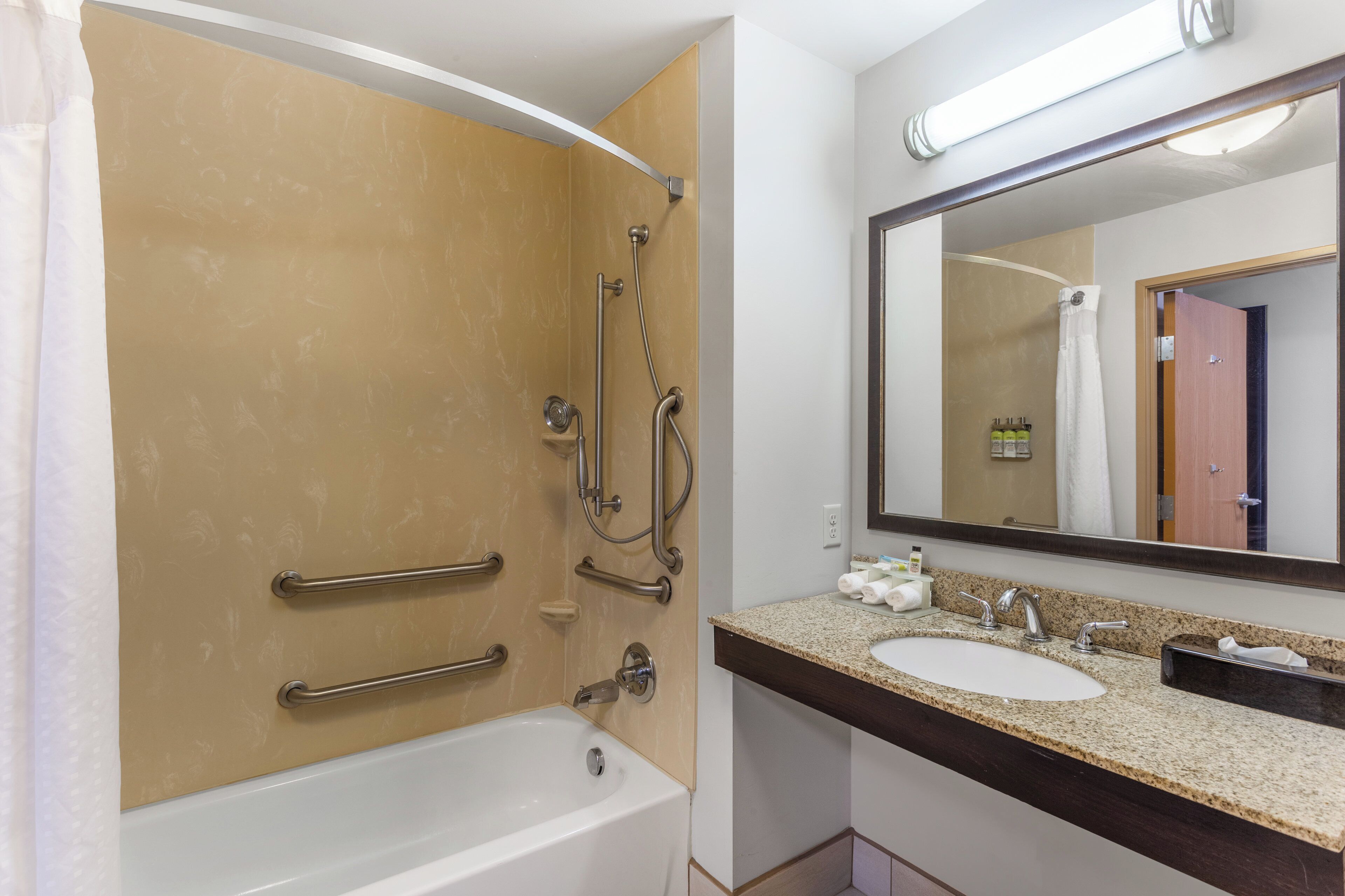 Un baño con bañera, ducha con barras de apoyo, lavamanos con espejo y artículos de aseo.