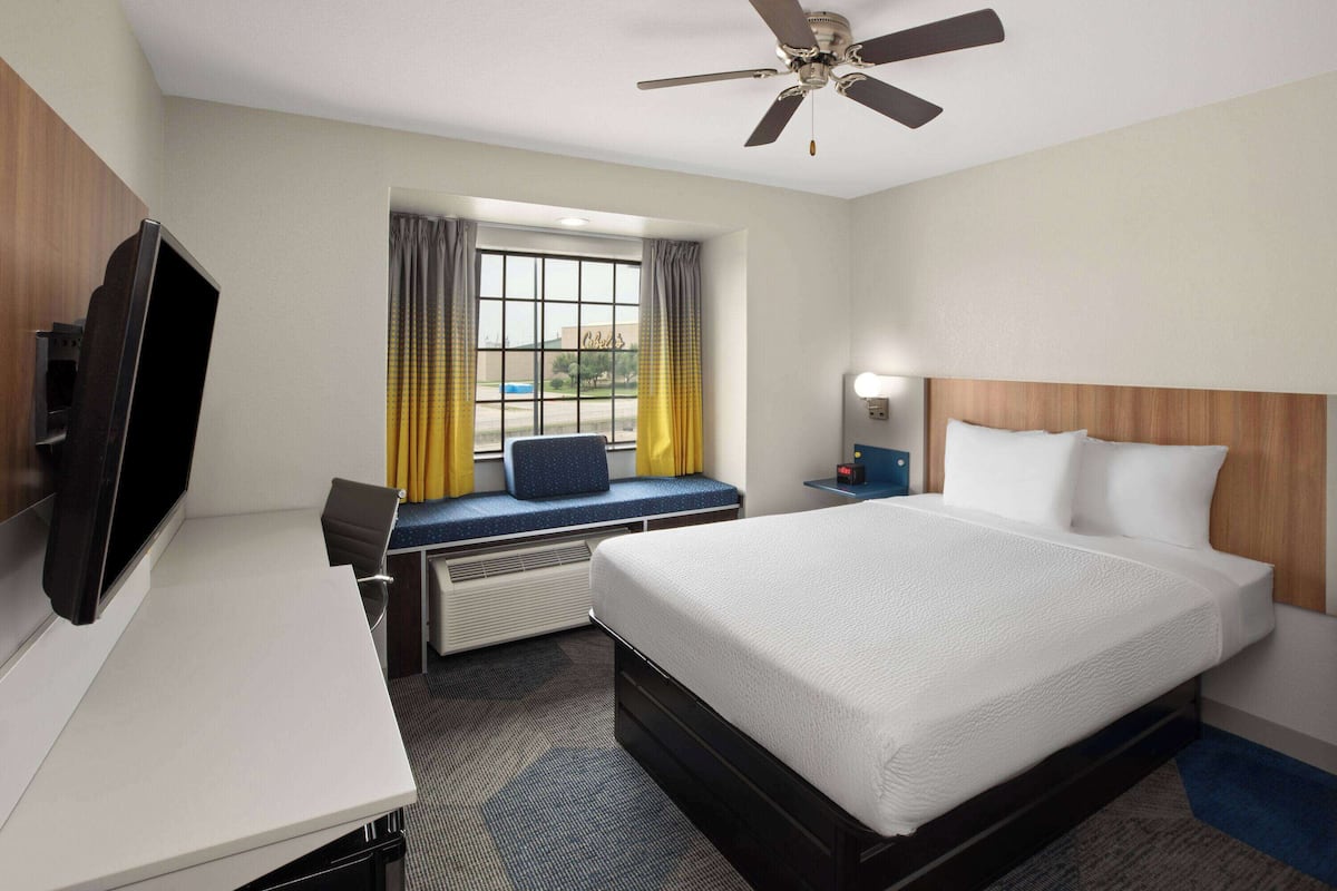 A hotel room with a bed, a desk with a TV, a window with curtains, a ceiling fan, and a nightstand with a lamp.