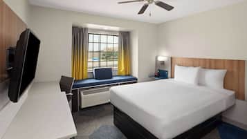 Una habitación de hotel con una cama, un escritorio con televisor, una ventana con cortinas, un ventilador de techo y una mesita de noche con lámpara.