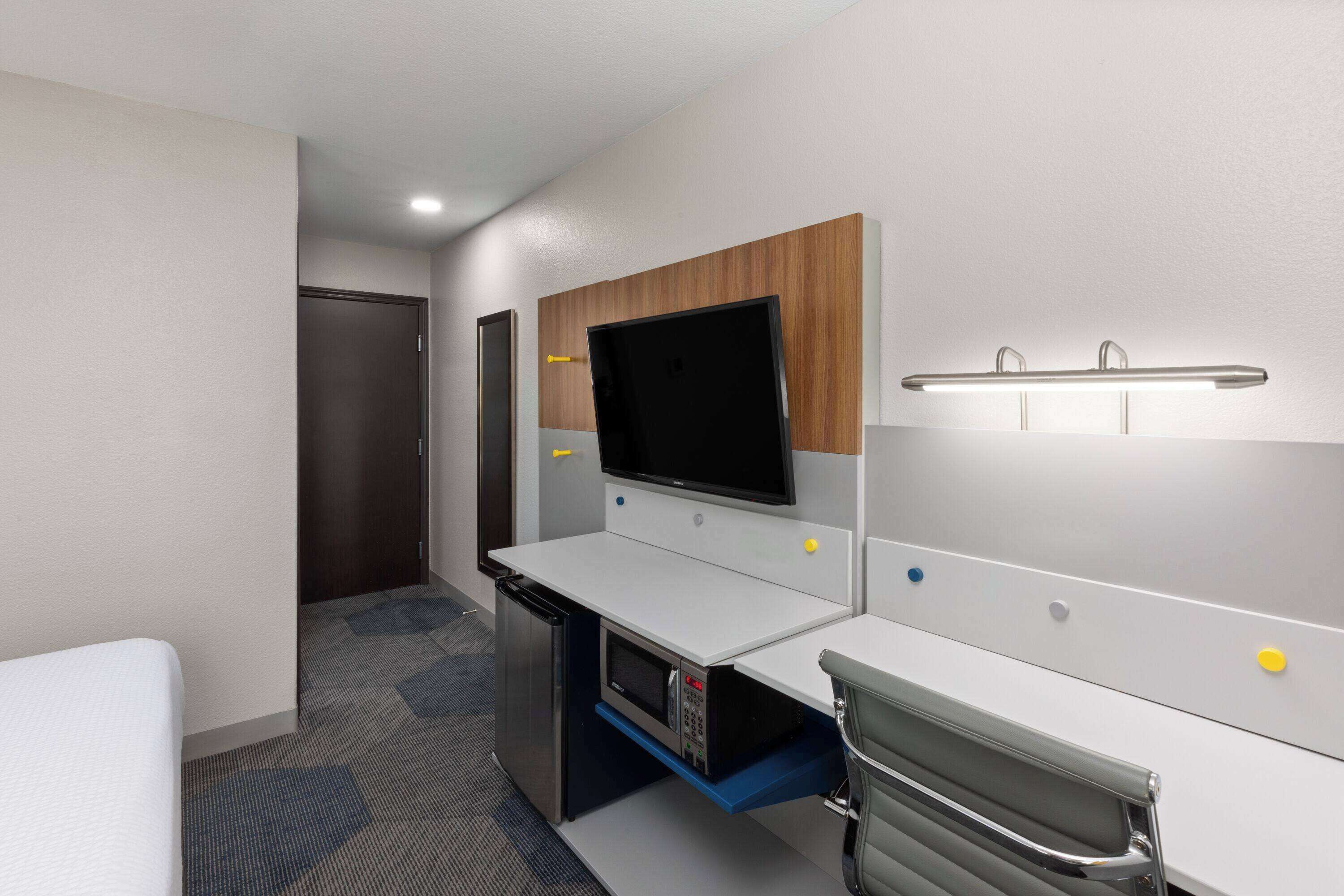 A hotel room with a bed, a desk with a computer, a TV, and a small kitchenette.