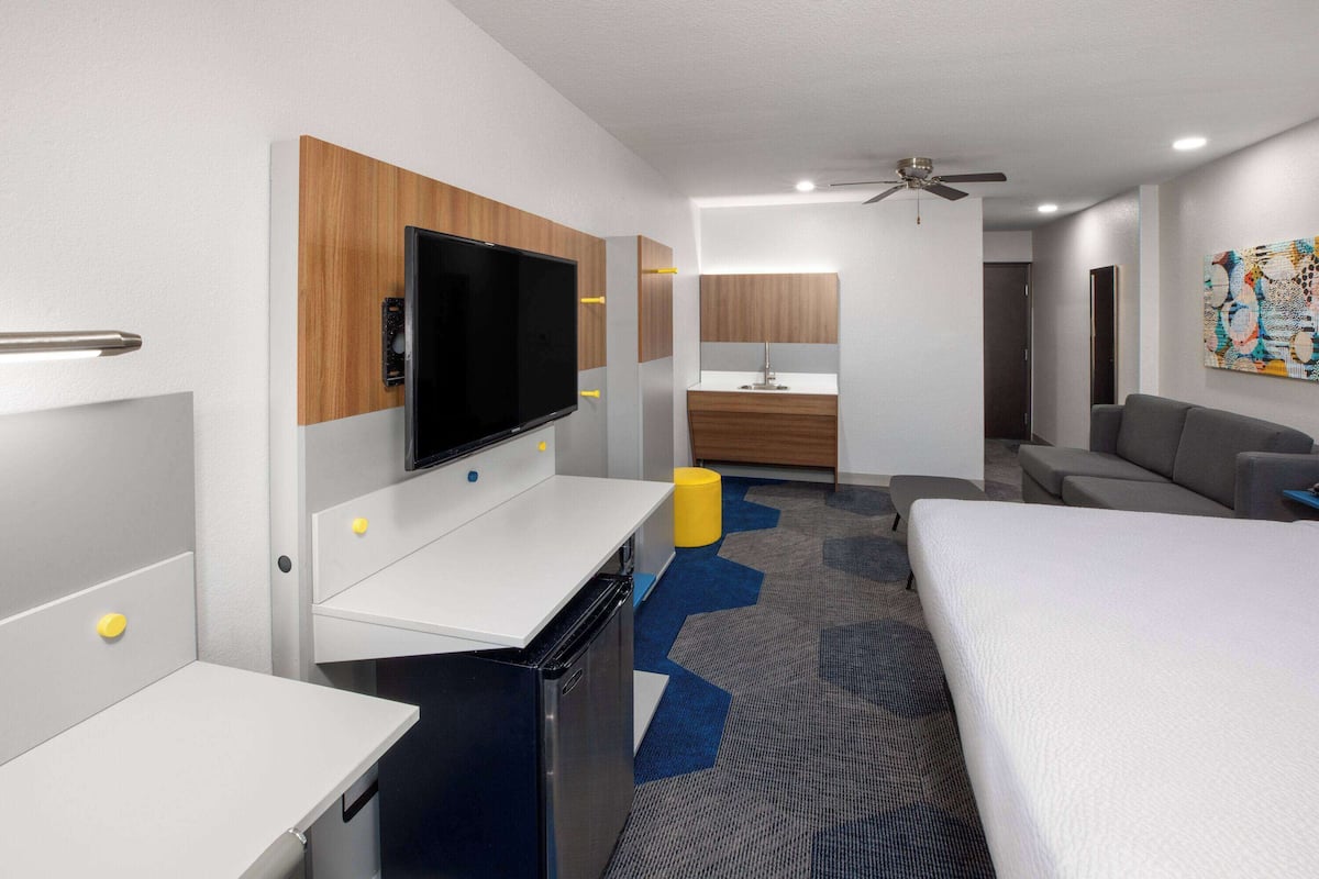 A hotel room with a bed, a desk with a TV, a small kitchenette, a sofa, and a ceiling fan.