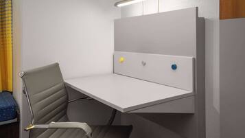 A compact, modern room with a built-in desk and chair, a small shelf, and a wall-mounted light fixture.